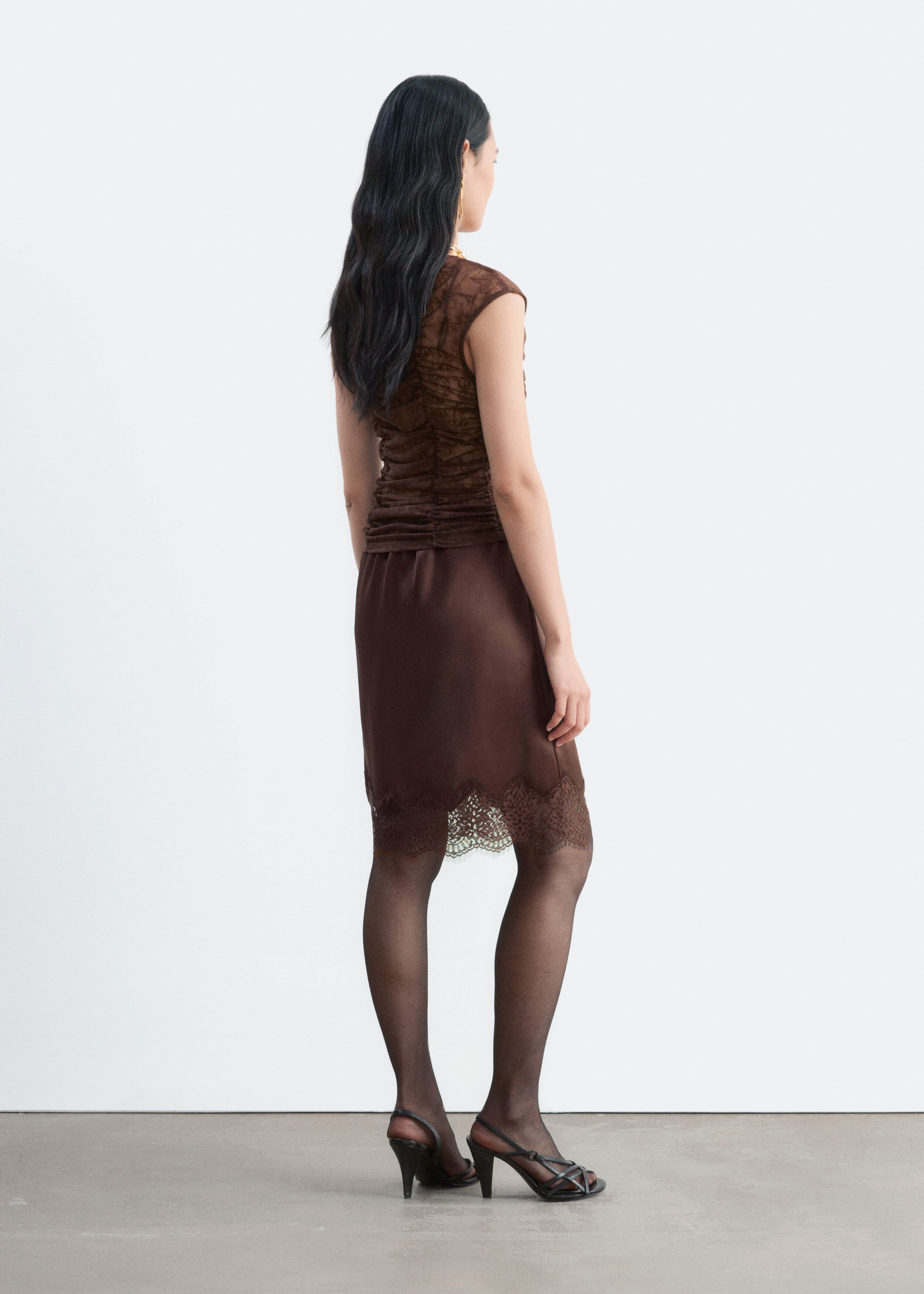 A brown lace top with ruching down the body and cap sleeves is being worn by the model, who is standing in a three-quarter rear view. The top is styled with a matching brown skirt featuring a lace-trimmed hem, sheer black tights, and black strappy heels.