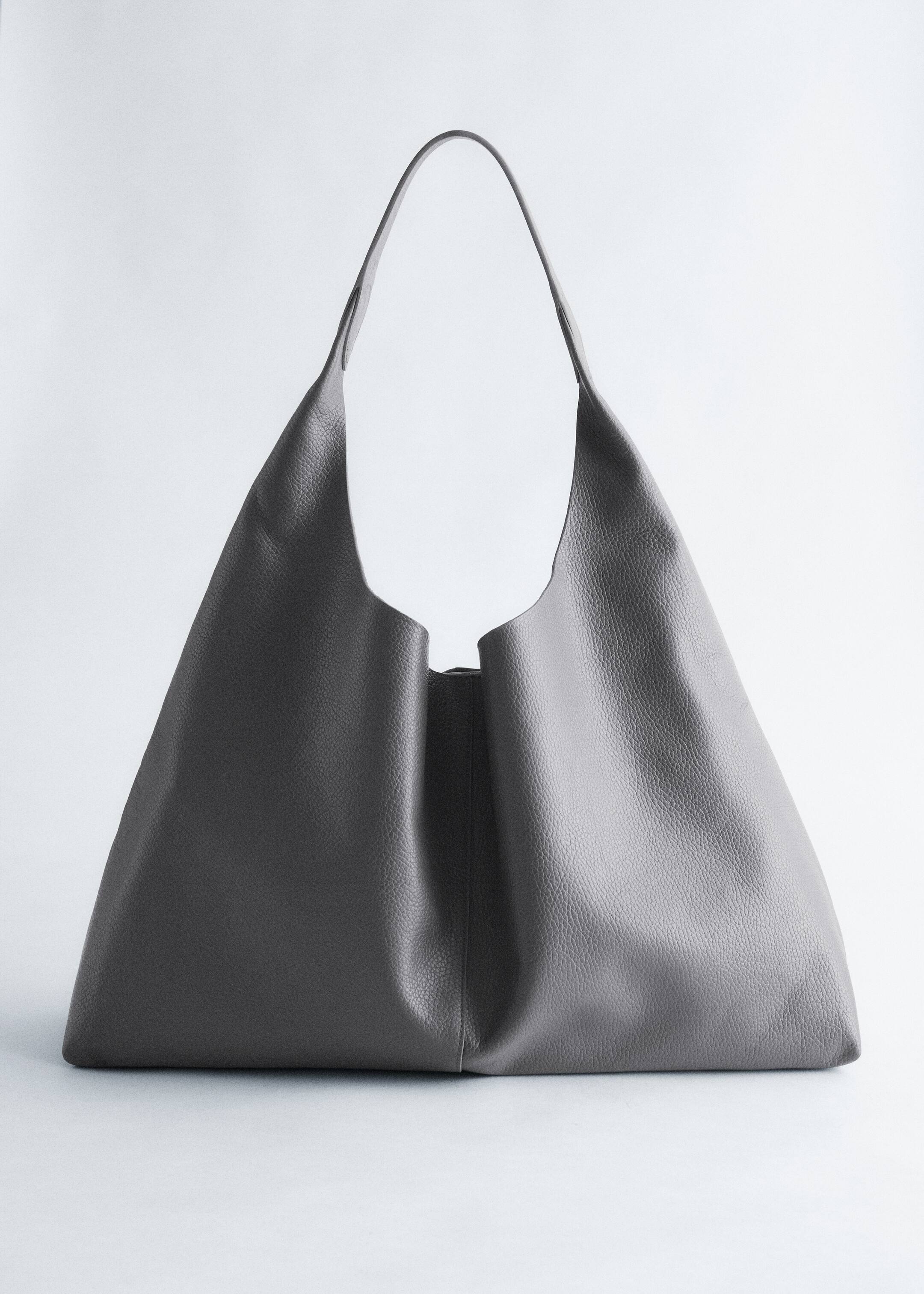 Agrandir l'image: Un sac à main gris foncé, à structure souple, est orienté vers l'avant, affichant sa texture granuleuse et une large base plate. Une seule et large sangle se courbe vers le haut depuis le corps du sac, et l'ouverture supérieure présente une subtile encoche en forme de V.