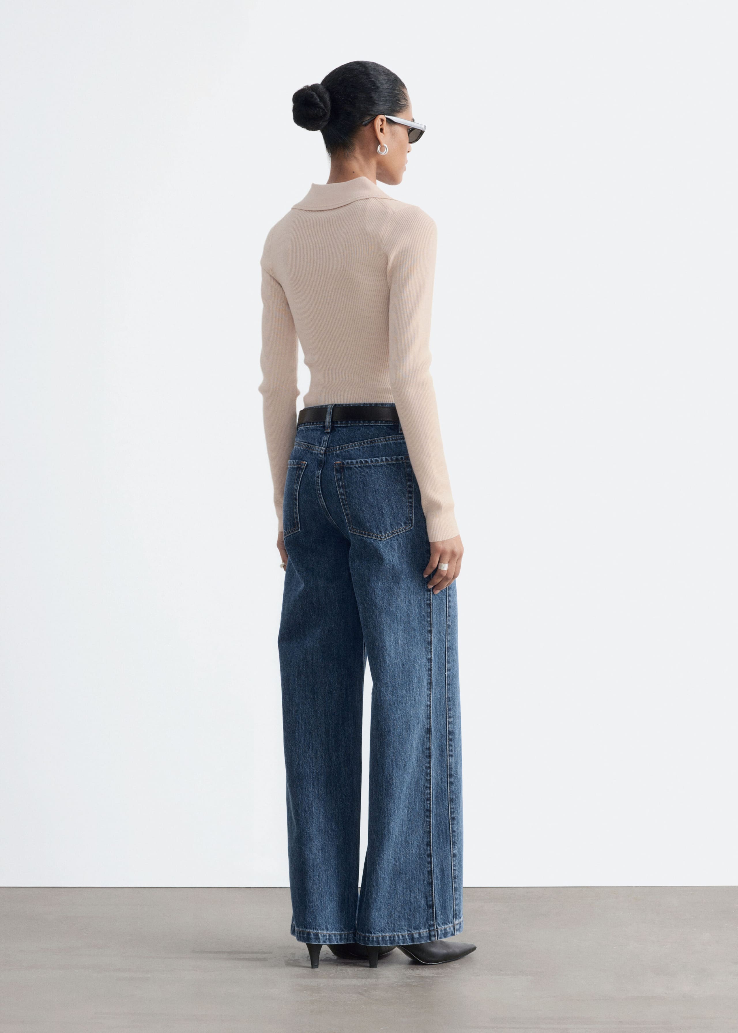 These high-waisted, wide-leg blue denim jeans are worn by the model and styled with a dark belt, a beige long-sleeve ribbed top, and black pointed-toe heels. Shown from a three-quarter rear angle, the jeans feature classic back patch pockets and a relaxed silhouette that falls to the heels.