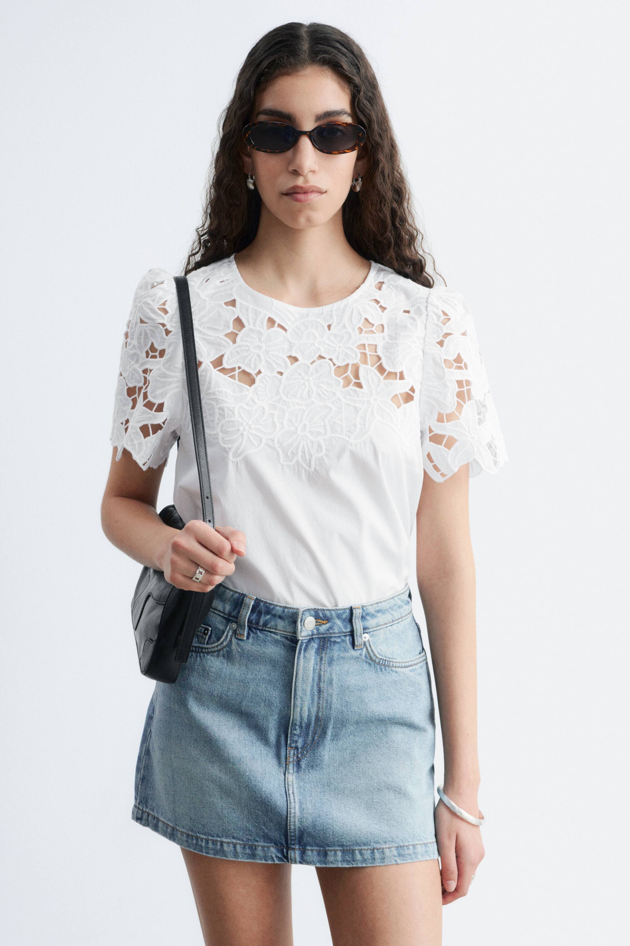 Agrandir l'image: Un haut blanc à manches courtes présente des découpes en dentelle à motifs floraux complexes sur le haut de la poitrine et les épaules, avec des manches légèrement bouffantes. Il est associé à une mini-jupe en denim bleu clair, qui se caractérise par une taille haute, des poches avant et un ourlet droit.