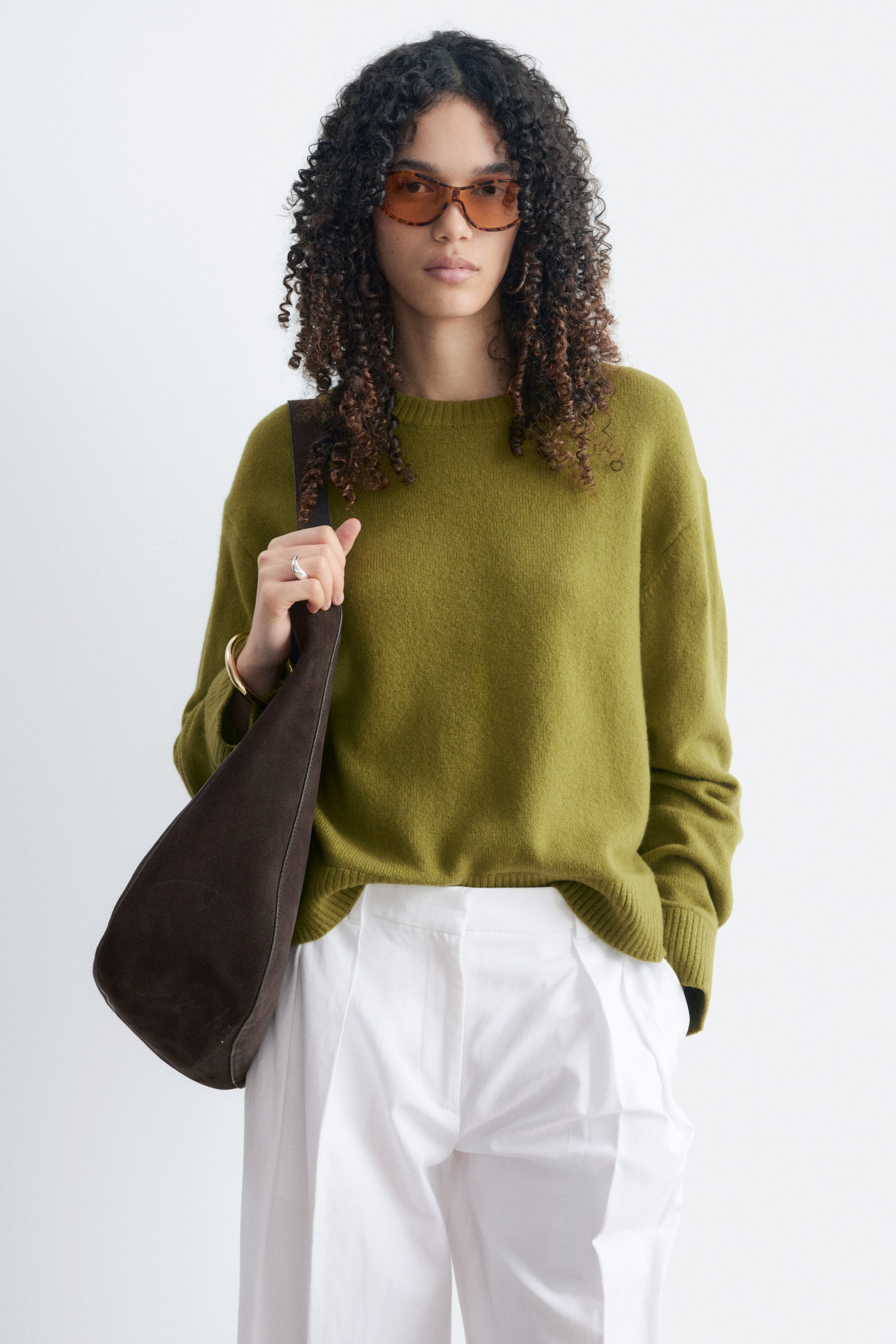 Agrandir l'image: Un pull en maille vert olive à coupe boxy, avec épaules tombantes et finitions côtelées, est porté par le mannequin. Un sac bandoulière marron foncé est porté par-dessus le pull, et celui-ci est stylisé avec un pantalon blanc.