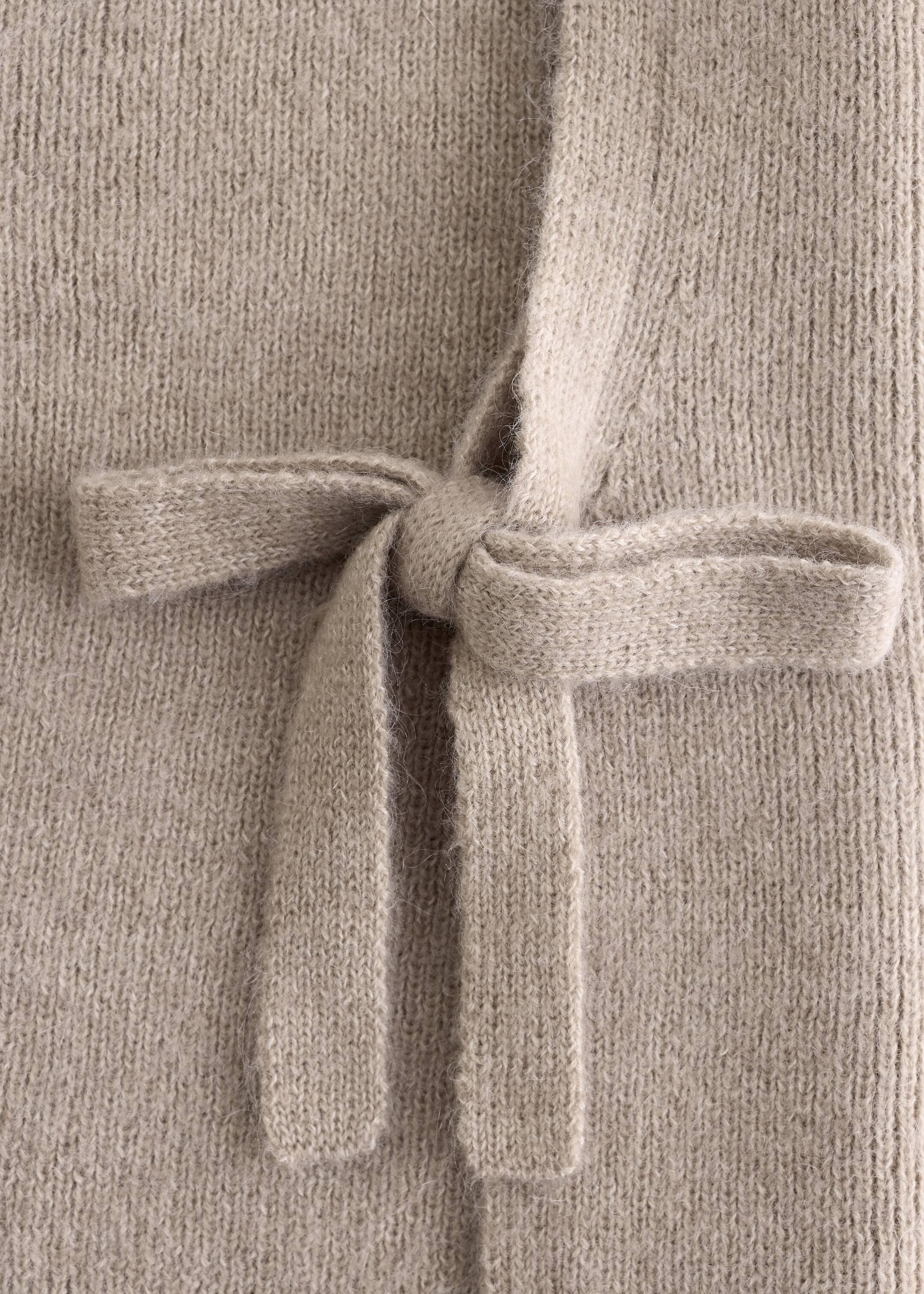 Grotere afbeelding bekijken: Een licht beige gebreide stropdas wordt in een strik geknoopt tegen een bijpassende gebreide achtergrond. De zachte, pluizige textuur van het breisel toont verticale rijen steken.