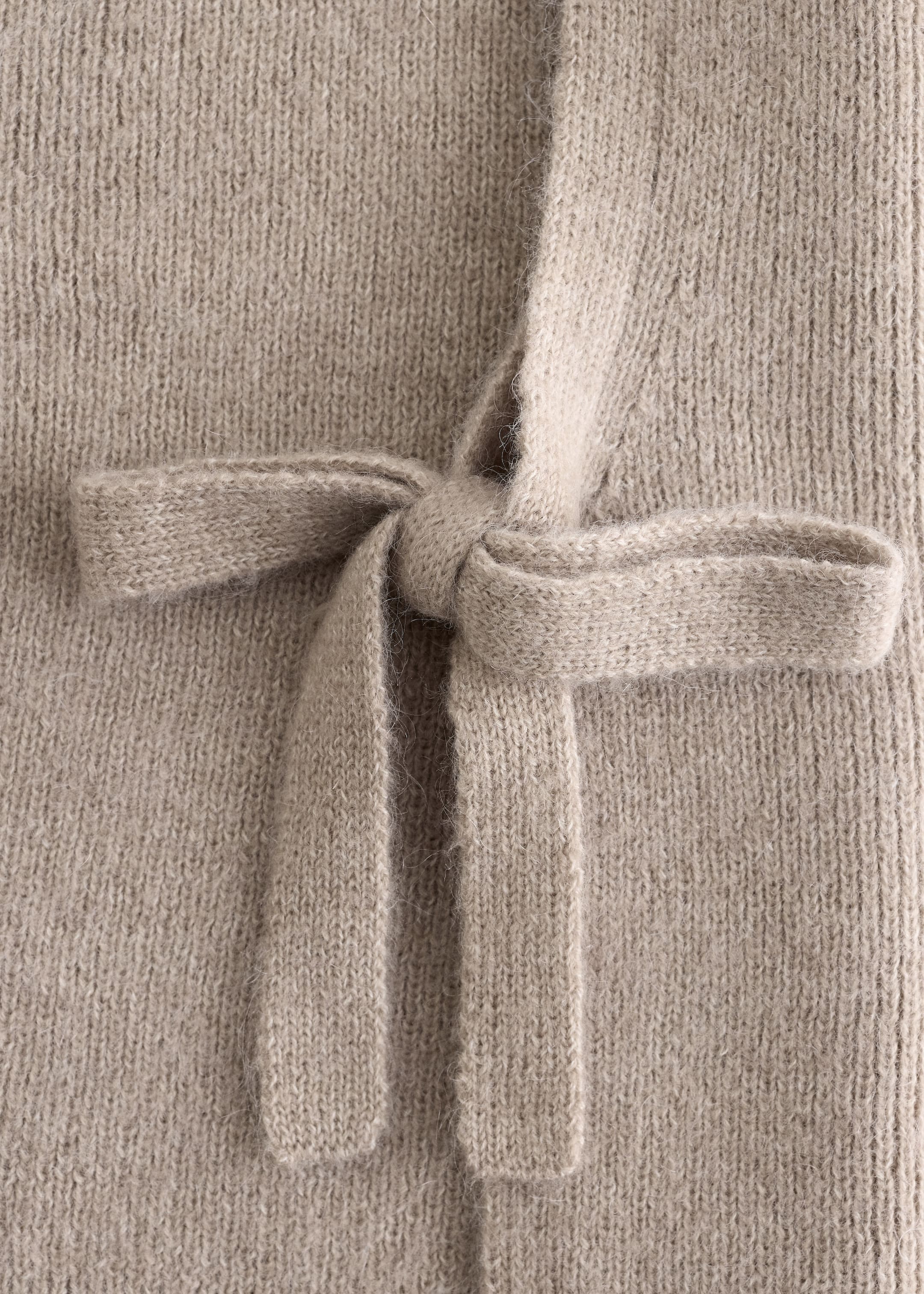 Visa större bild: En ljusbeige stickad slips knyts till en rosett mot en matchande stickad bakgrund. Den mjuka, luddiga texturen av stickningen har vertikala rader av stygn.