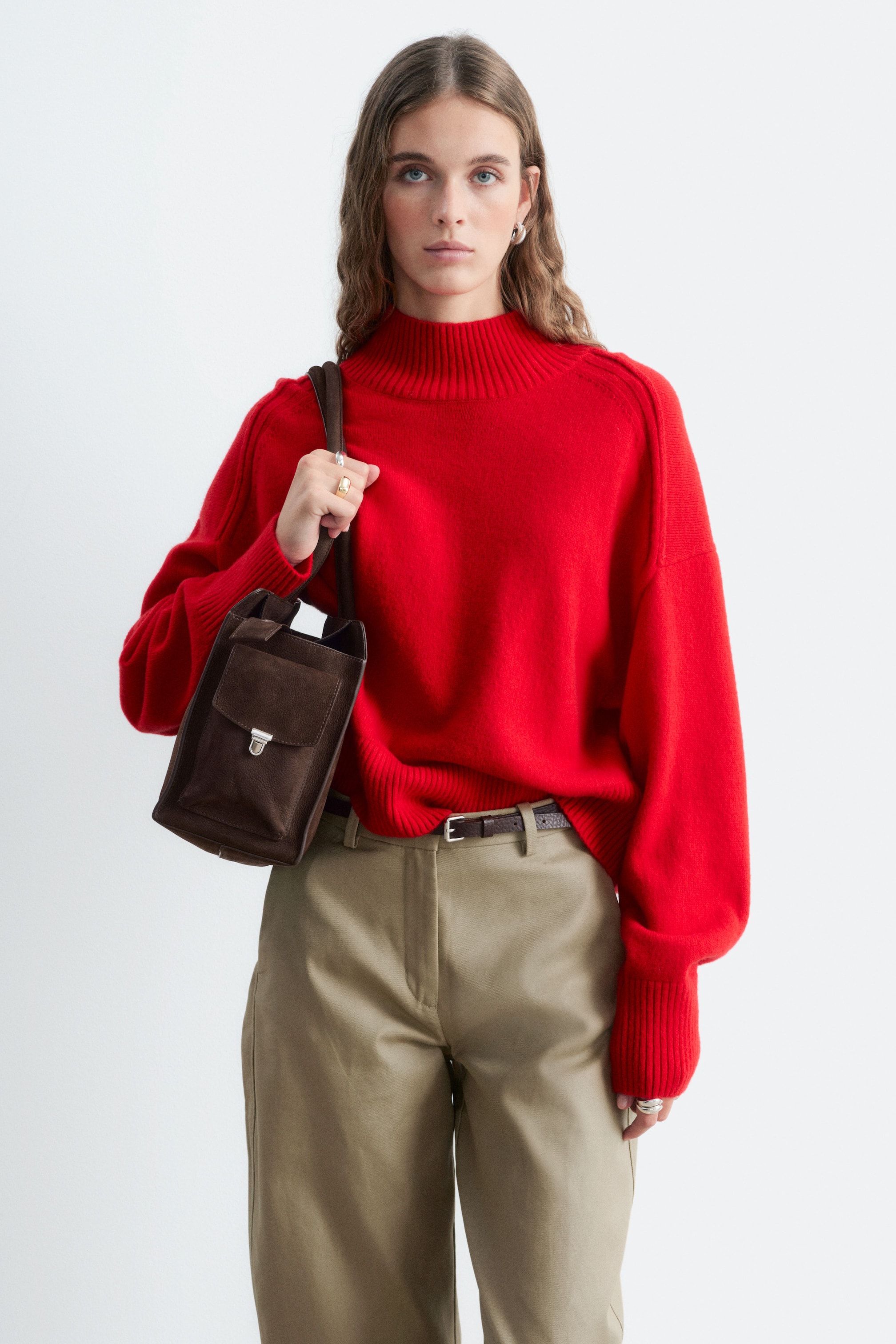 Agrandir l'image: Un pull col montant rouge à coupe décontractée, avec emmanchures tombantes et détails côtelés au col et aux poignets, est porté par le mannequin. Le pull est associé à un pantalon beige et à un sac bandoulière en daim marron foncé que le mannequin tient à la main.