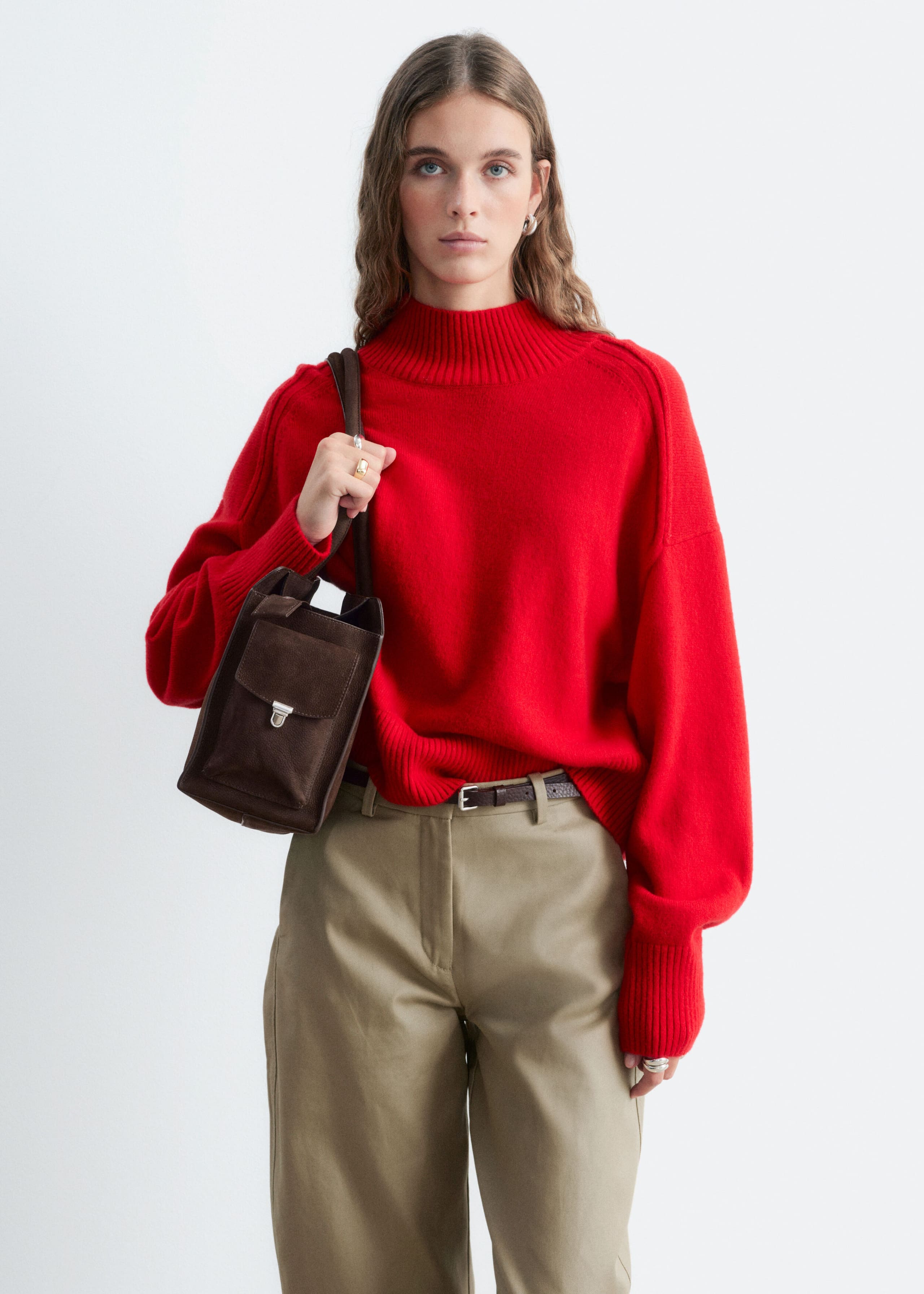 A red mock-neck sweater with a relaxed fit, dropped shoulder seams, and ribbed detailing on the collar and cuffs is worn by the model. The sweater is styled with beige trousers and a dark brown suede shoulder bag that the model is holding.