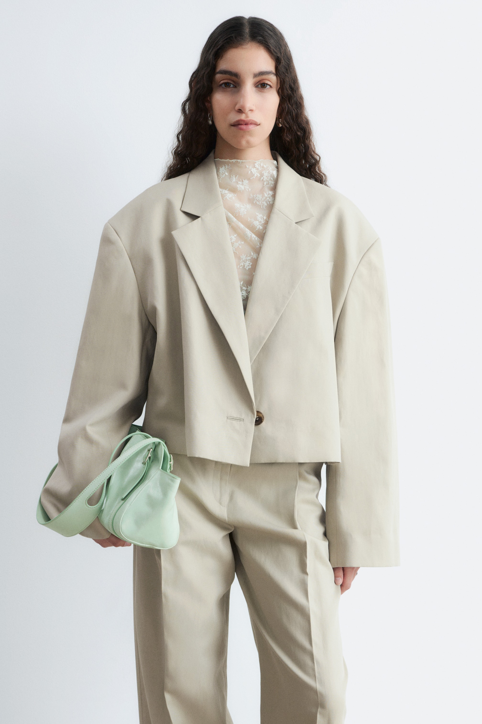 View larger image: Presenting a light beige cropped blazer paired with matching wide-leg trousers. The blazer showcases oversized shoulders, notch lapels, and a single brown button fastening, with a sheer floral top visible beneath.