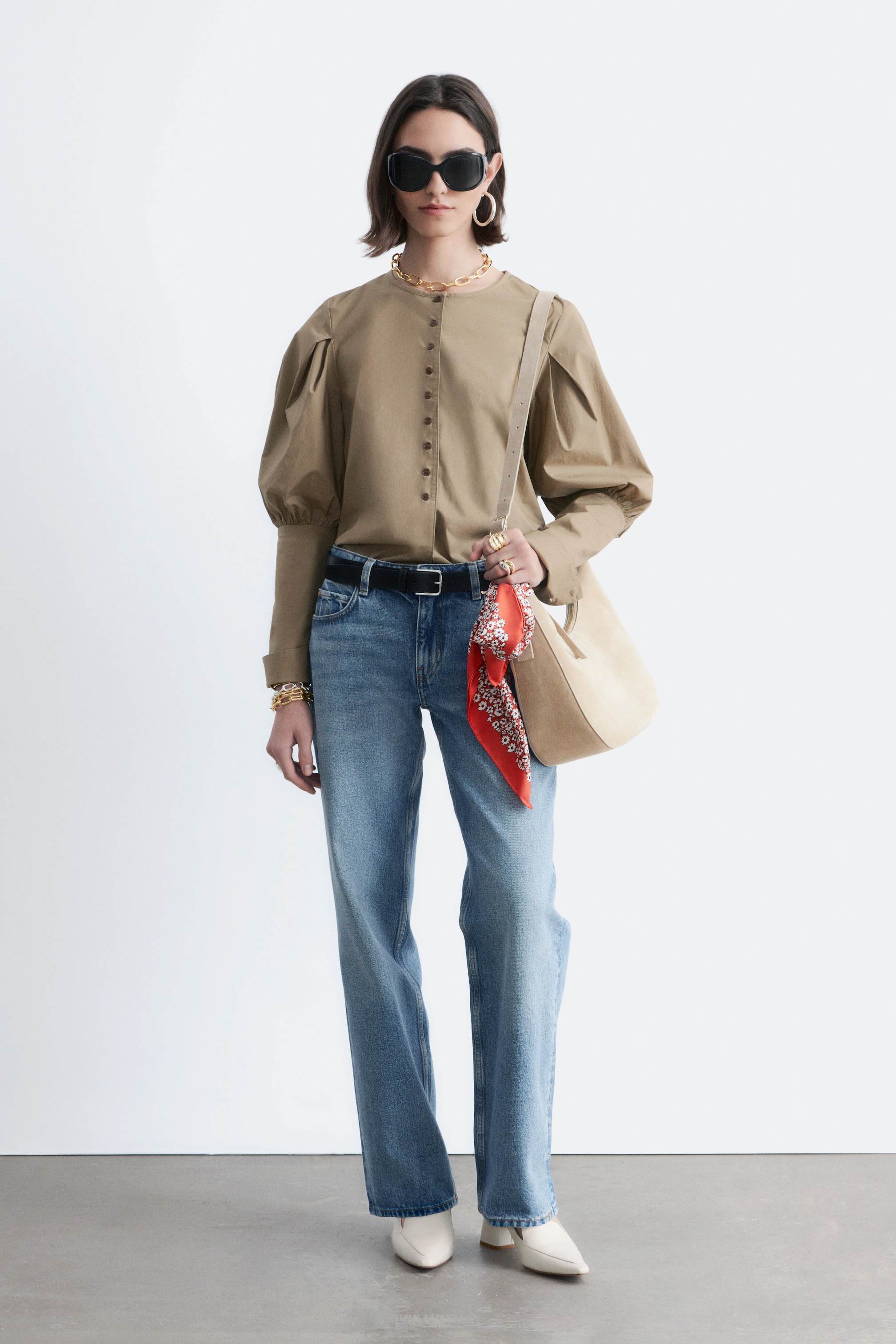 Ver imagen más grande: Una blusa marrón caqui se presenta de frente, con un escote redondo sin cuello y una tapeta completa de botones pequeños. Está diseñada con mangas largas abullonadas recogidas en los hombros, terminando en puños ajustados, y aparece metida dentro de los pantalones.