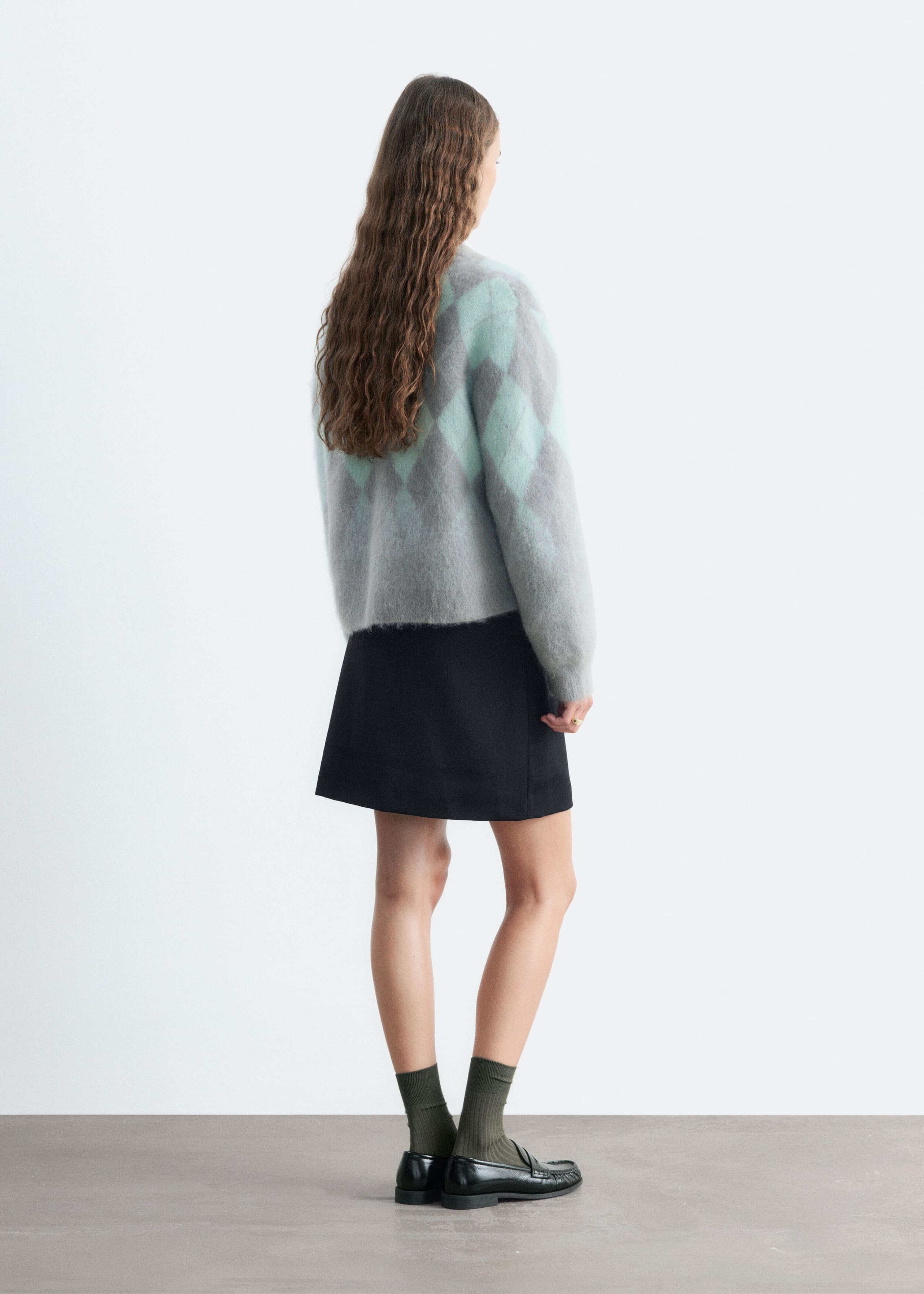 A mohair-blend argyle cardigan featuring a gray and light green diamond pattern is worn by the model, highlighting its fuzzy texture from the back. The long-sleeve cardigan is styled with a short black skirt, dark green socks, and black loafers.