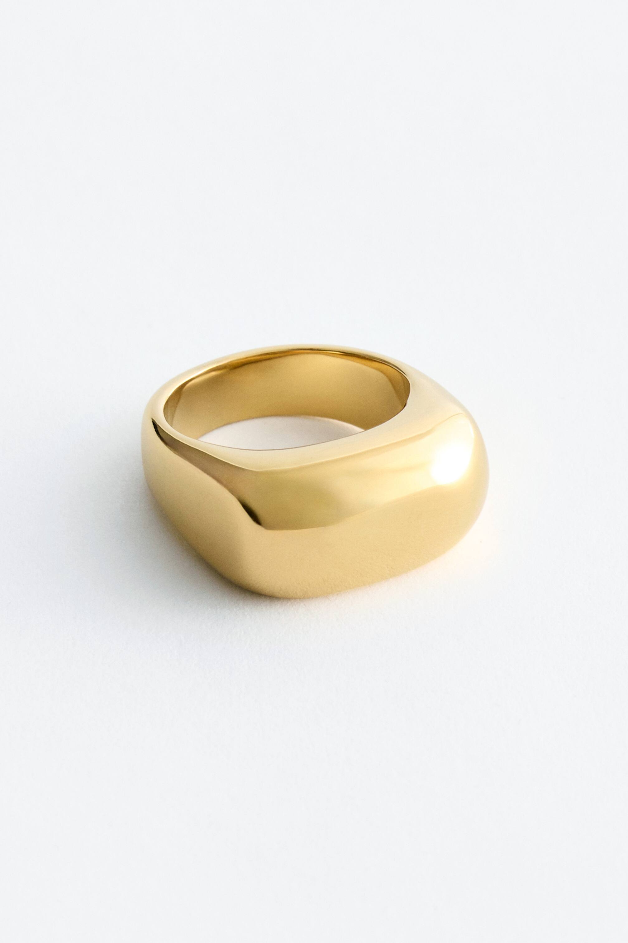 Grotere afbeelding bekijken: Een gouden ring met een dikke, gepolijste band en een subtiel vierkant, afgerond bovenvlak, lichtjes gekanteld om de binnenomtrek te onthullen.