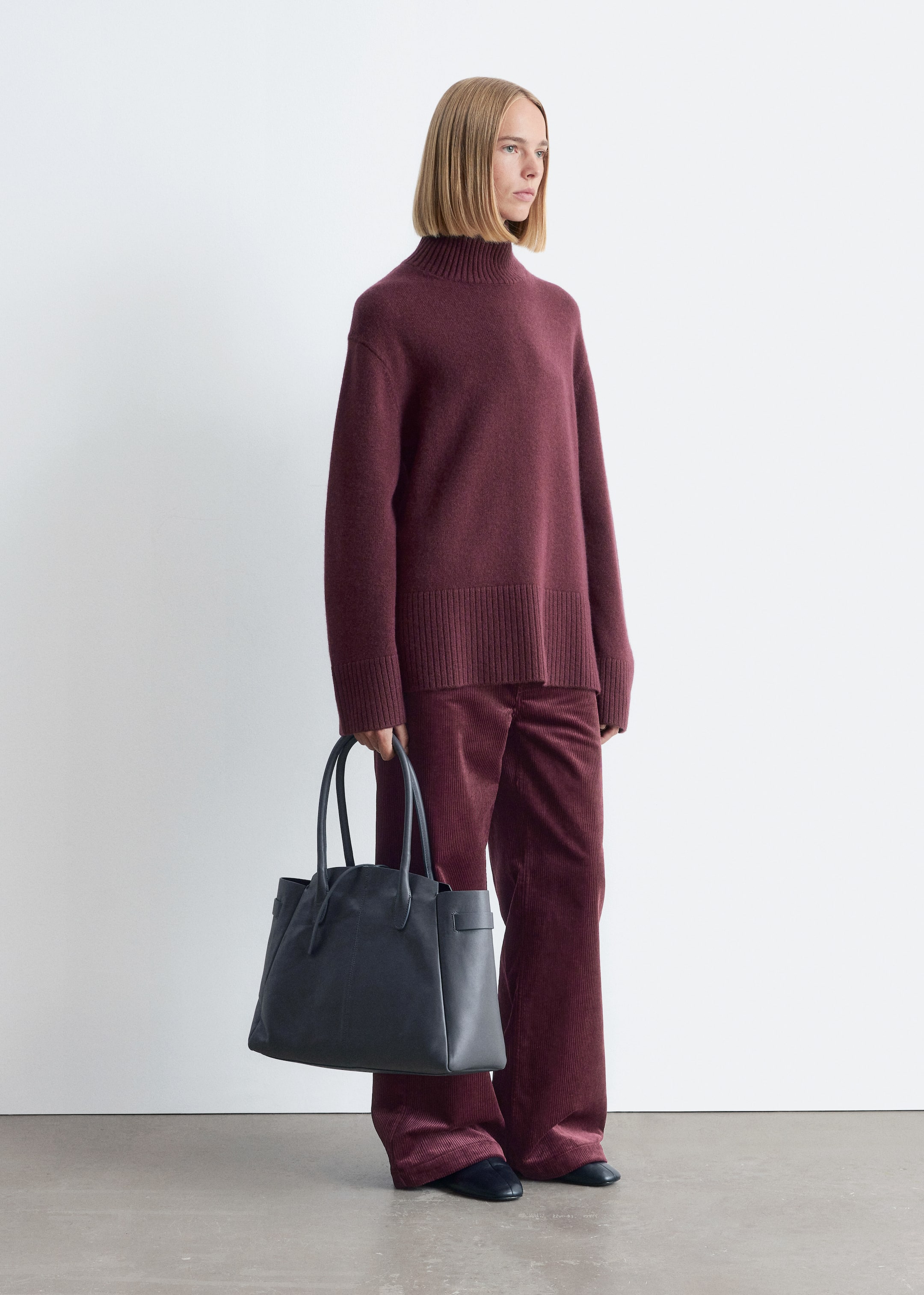 Grotere afbeelding bekijken: De model draagt een oversized donkerrode coltrui met een geribbelde kraag en zoom, samen met bijpassende wijde corduroy broek en houdt een donkergrijze draagtas vast.