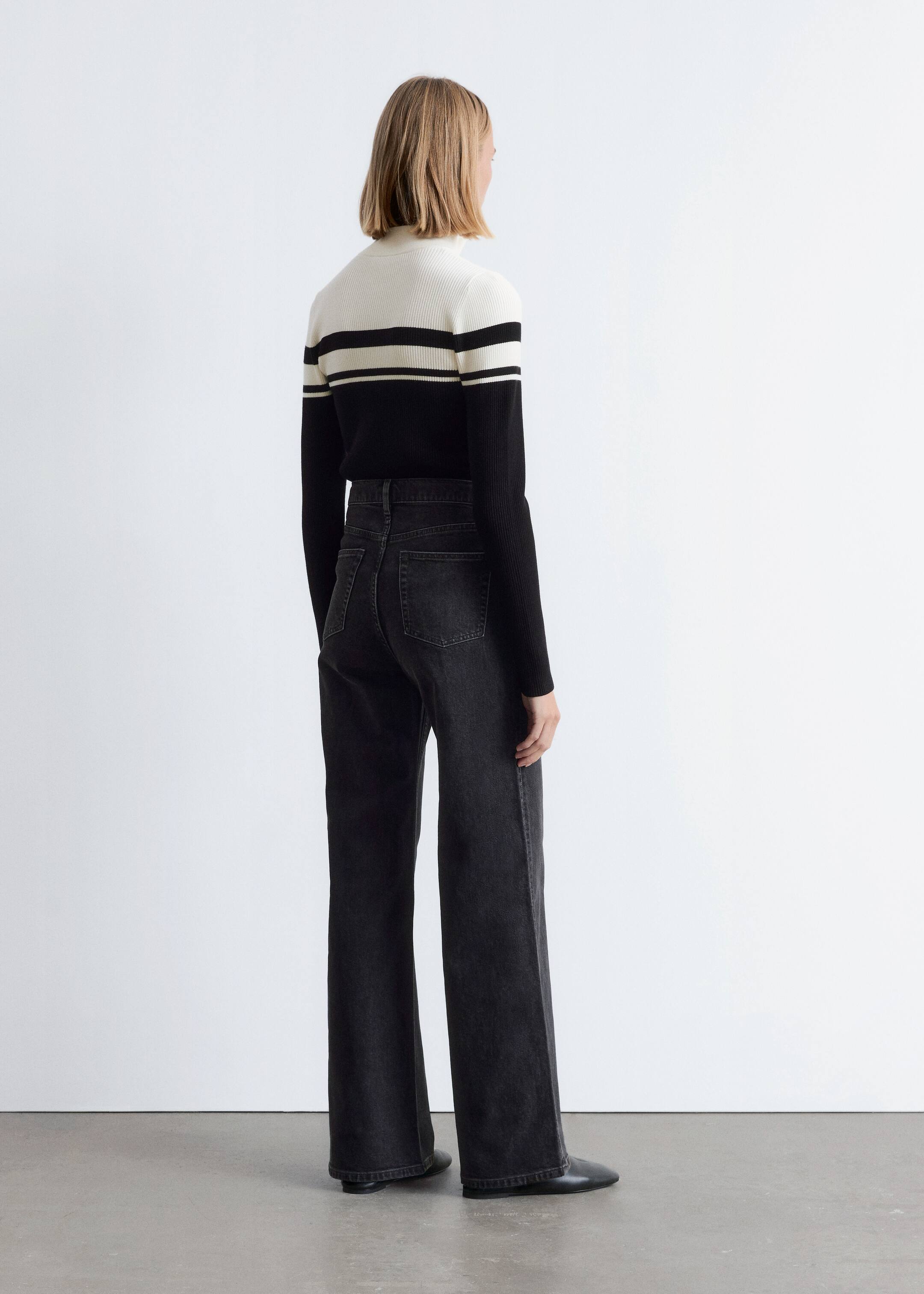 View larger image: A black and white ribbed turtleneck sweater is being worn, featuring a white upper body, black lower body, and two thin white horizontal stripes across the chest. It is paired with high-waisted, wide-leg dark gray jeans, viewed from a back-right angle.