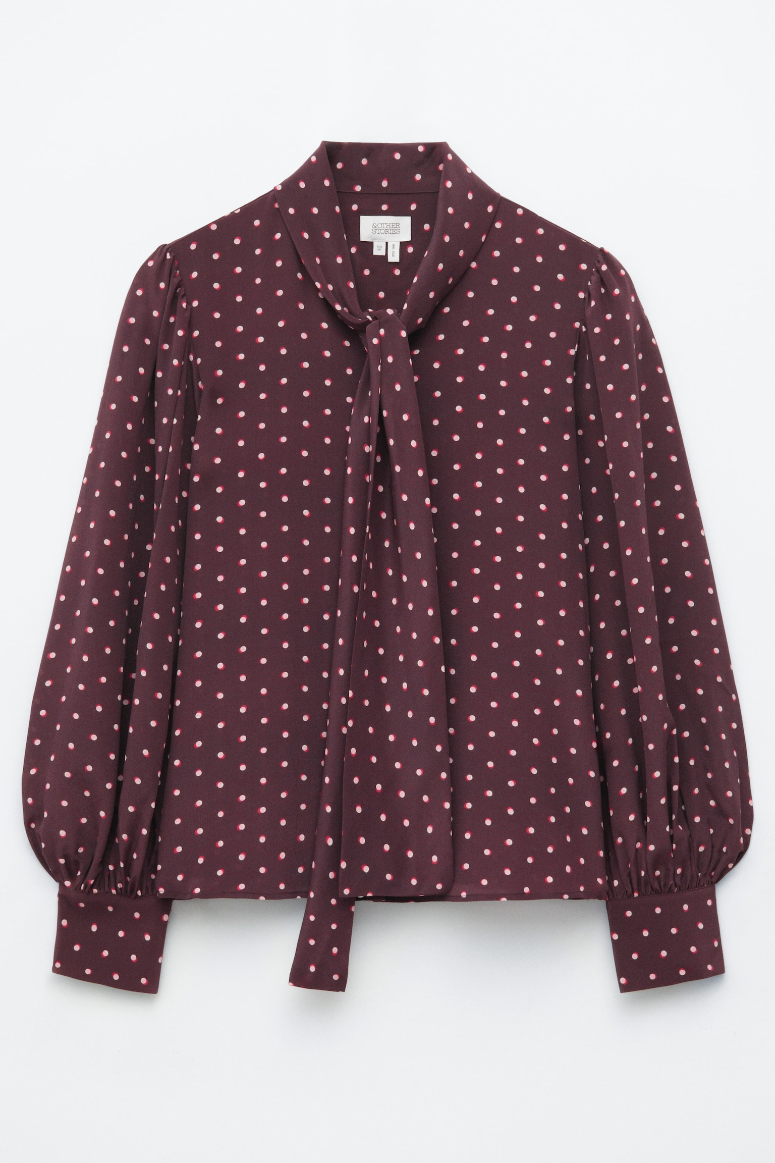 Una blusa in seta bordeaux con stampa a pois all-over è vista frontalmente e mette in evidenza un colletto alto con una sciarpa a doppio strato applicata, annodata al centro. La blusa presenta maniche lunghe con spalle arricciate e polsini ampi con bottoni.
