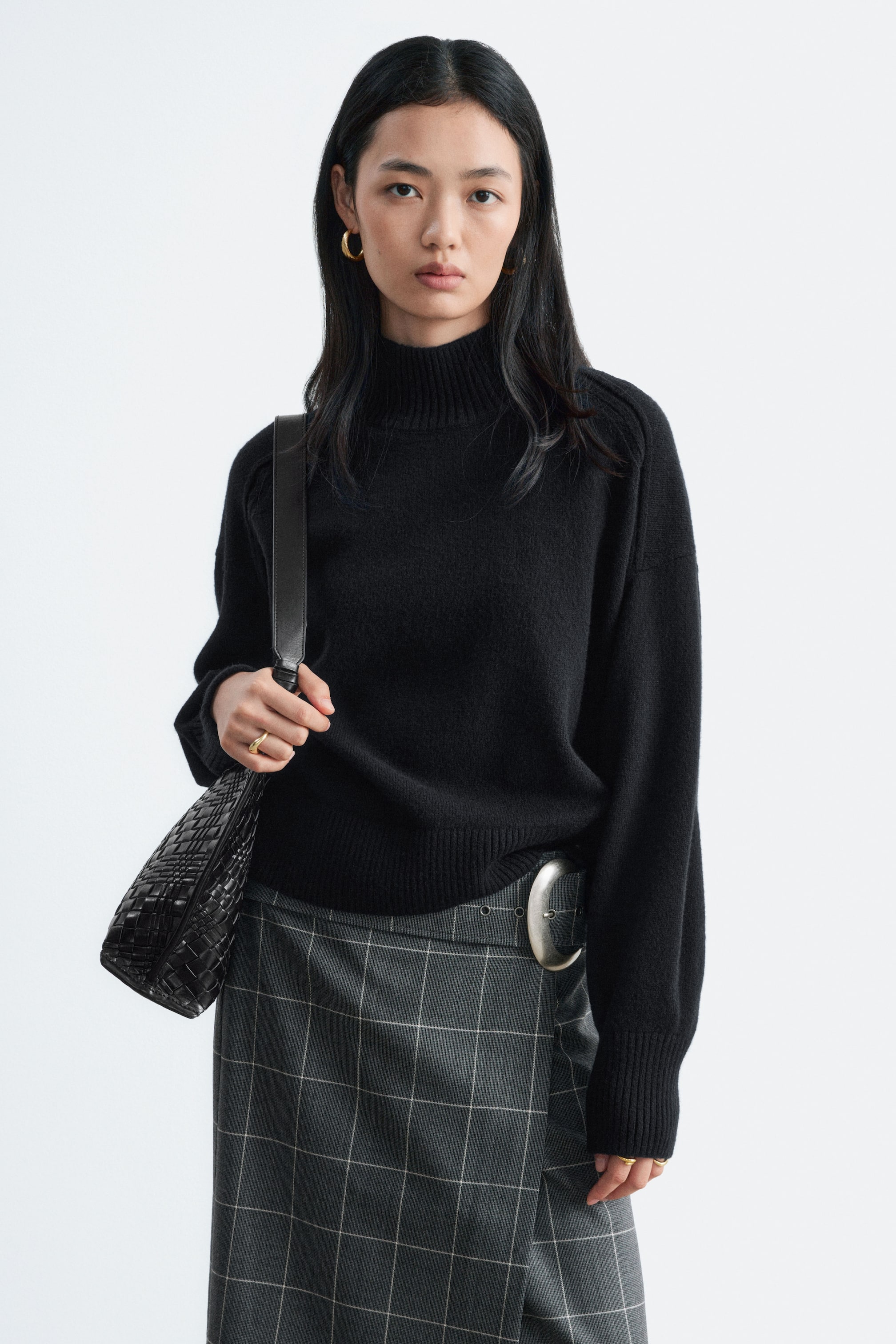 Größeres Bild ansehen: Ein schwarzer Pullover mit Stehkragen und Rippbündchen sowie legeren Schultern wird vom Model getragen. Der Pullover ist mit einem grauen Wickelrock mit Karomuster und großer Schnalle kombiniert, und das Model hält den Riemen einer schwarzen gewebten Schultertasche.