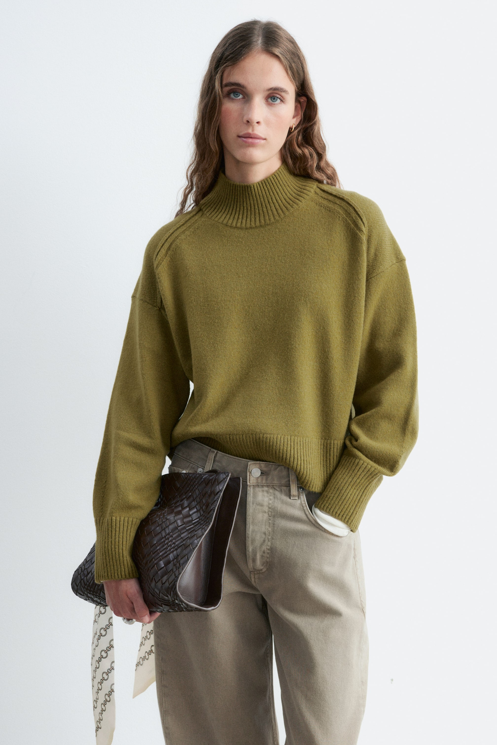 Größeres Bild ansehen: Ein olivgrüner Pullover mit Stehkragen und Rippbündchen an Kragen, Ärmeln und Saum wird vom Model getragen. Das Model kombiniert den Pullover mit einer beigen Hose und hält unter einem Arm eine dunkelbraune, geflochtene Handtasche, während es die andere Hand in der Tasche ruhen lässt.
