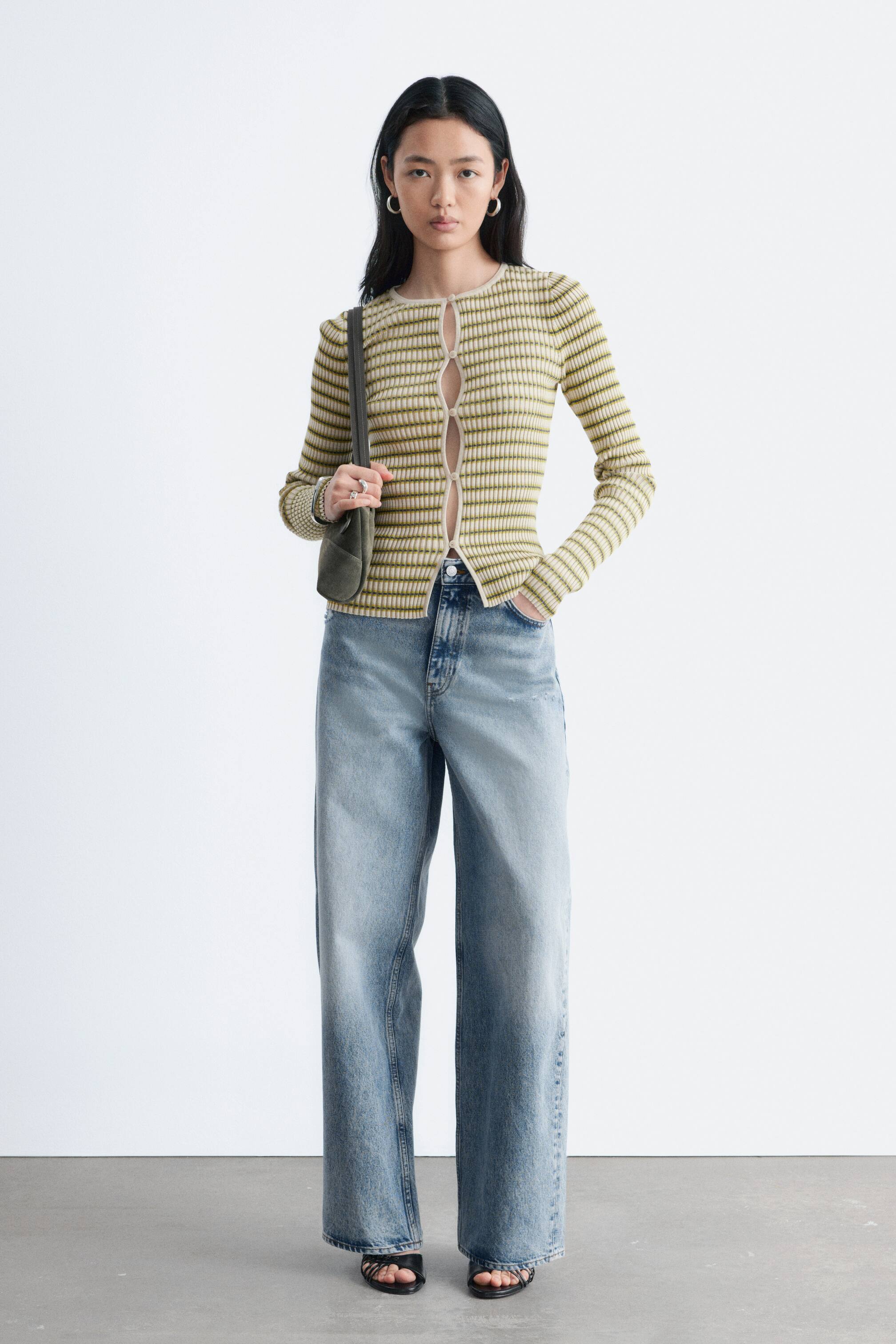 Ingrandisci l'immagine: Il cardigan è un modello aderente e corto che presenta strisce orizzontali verdi e bianco sporco in maglia a coste. È progettato con un collo rotondo e maniche lunghe, e la chiusura con bottoni sul davanti mostra i primi due bottoni abbottonati mentre i bottoni inferiori sono sbottonati.