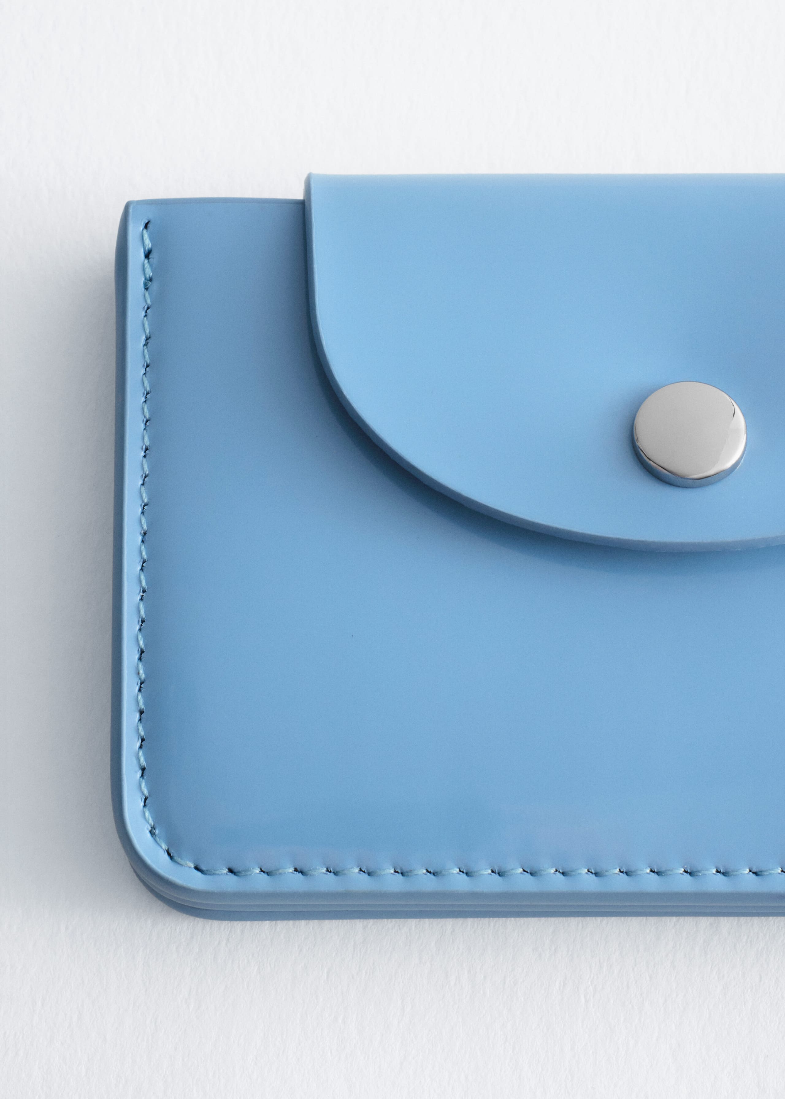 A light blue card holder is shown in a close-up front view, showcasing a curved top flap with a silver-tone snap closure and tonal stitching along its rounded edges.