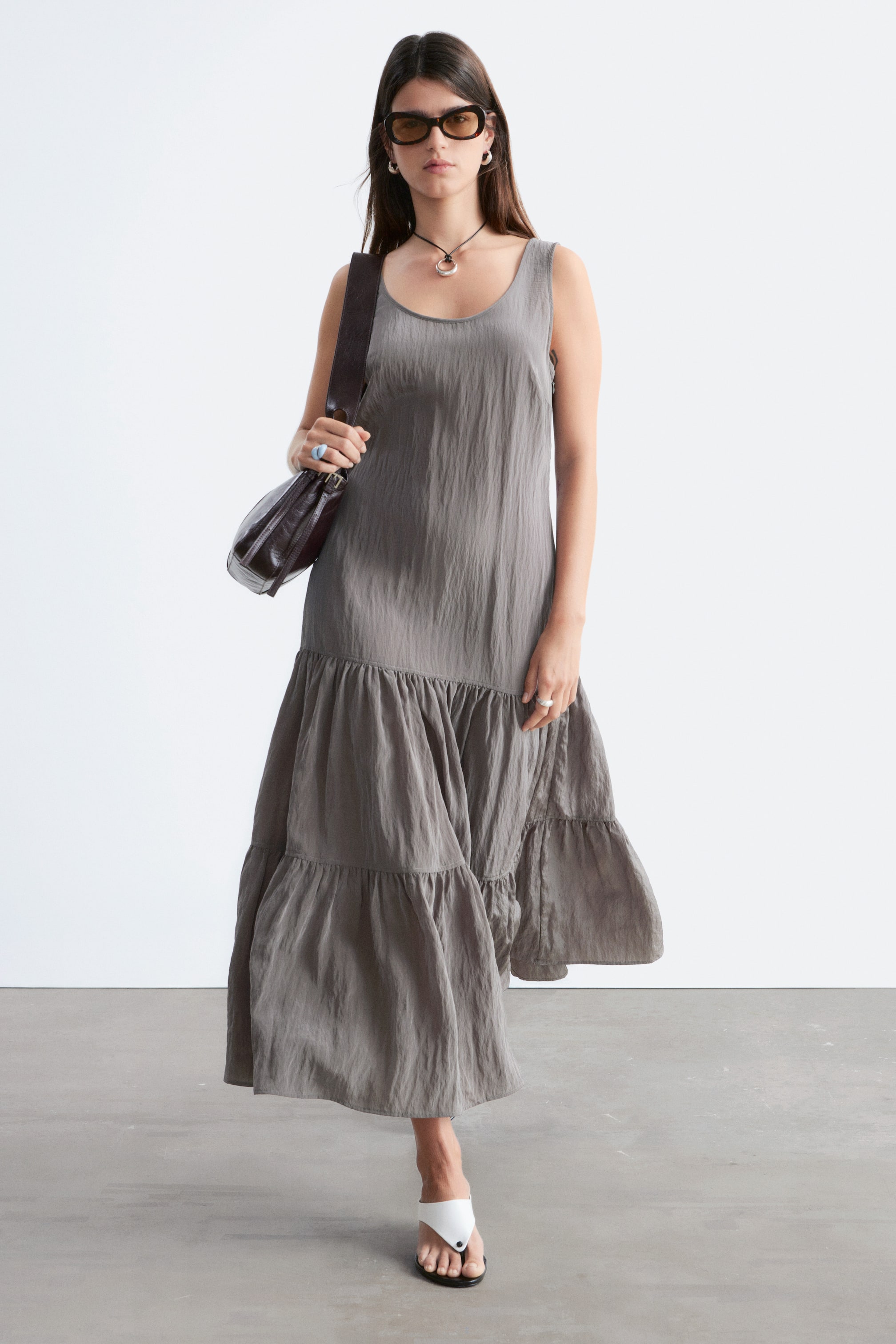 Agrandir l'image: Une robe maxi grise sans manches présente un décolleté rond et une texture froissée. Sa longue jupe comporte plusieurs volants froncés, créant une silhouette fluide.