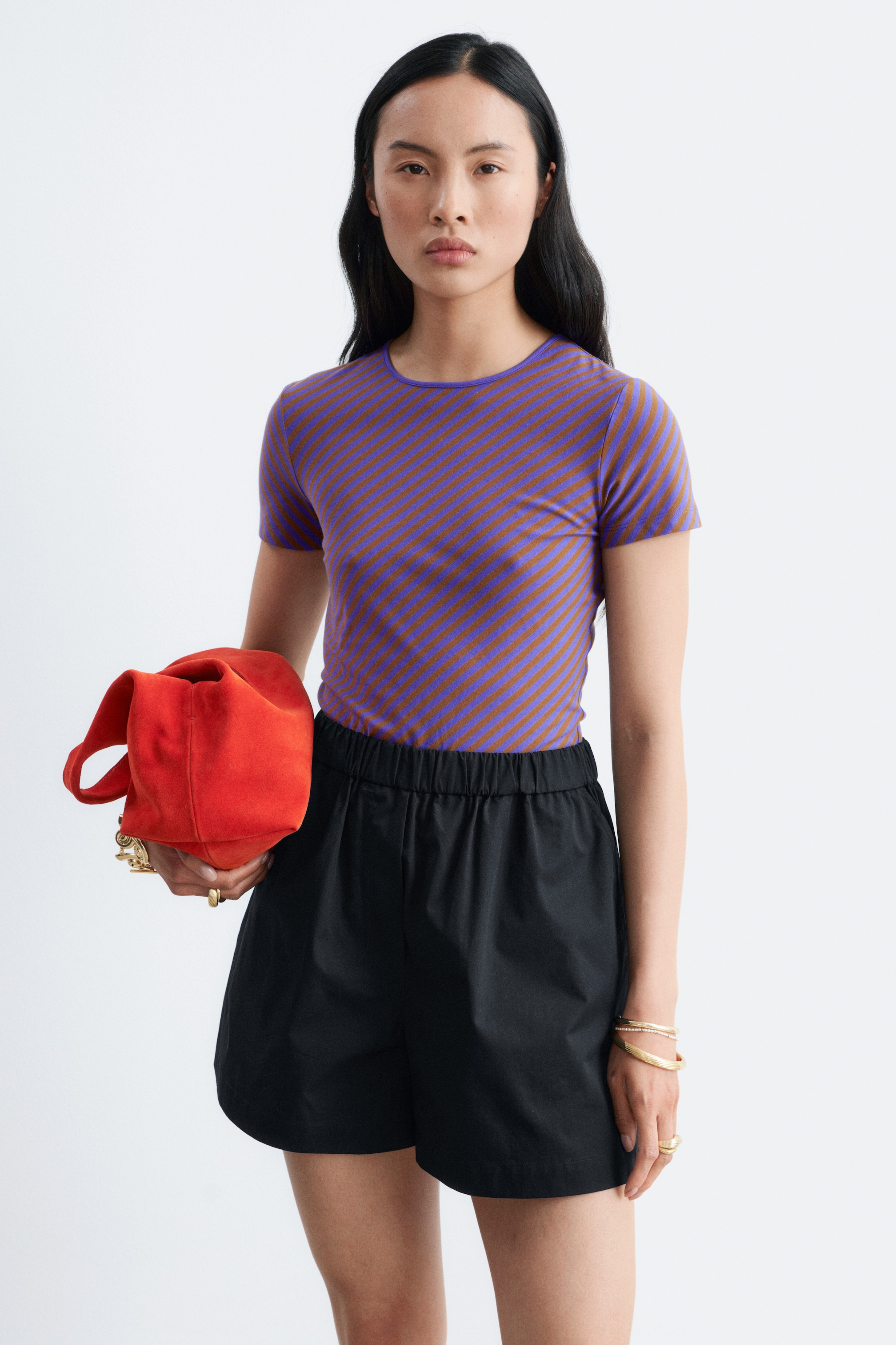 Visualizza immagine più grande: La modella indossa una maglietta in cotone sottile con un girocollo a coste e strisce diagonali viola e marroni. La t-shirt a maniche corte è abbinata a pantaloncini neri.