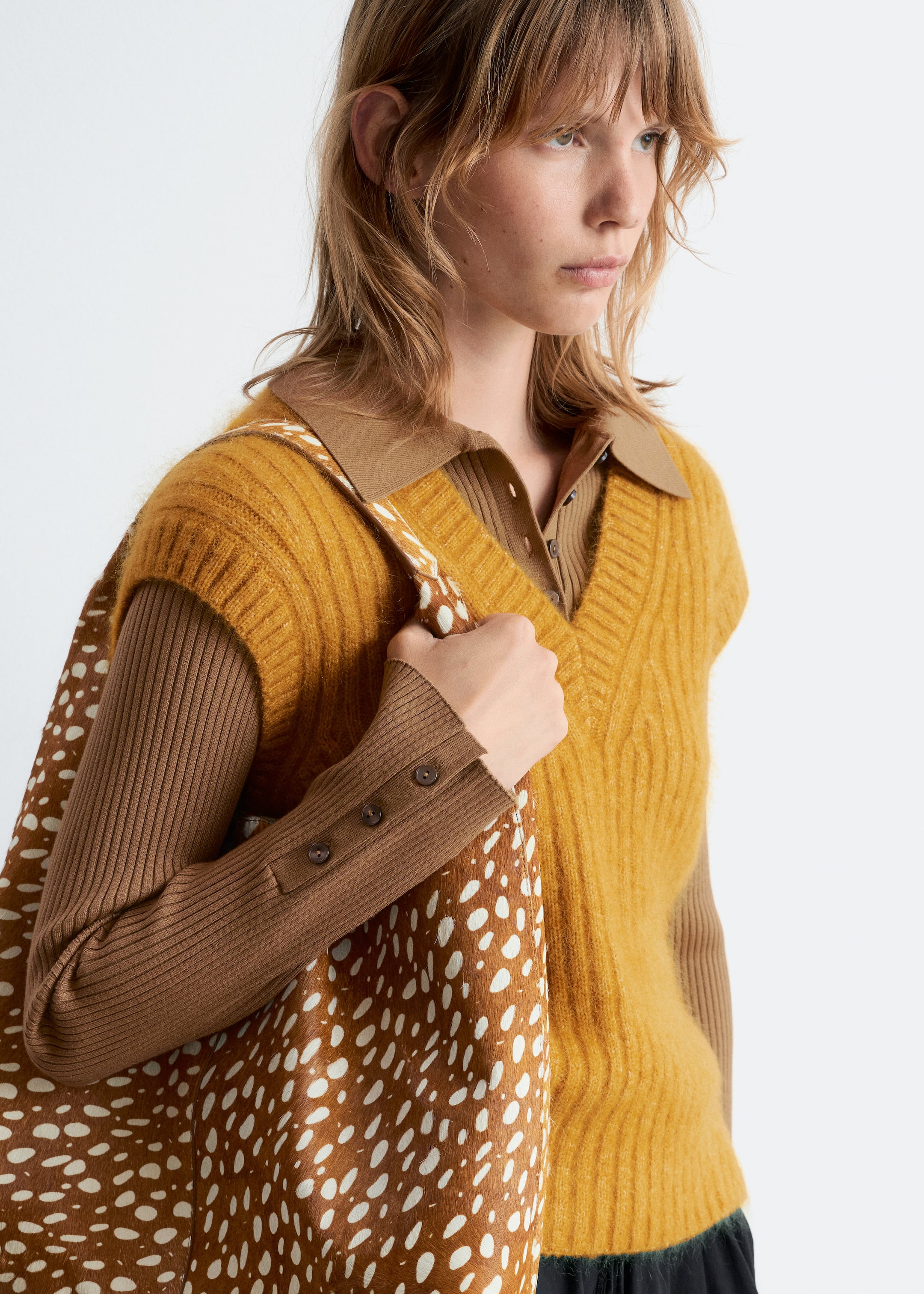 Agrandir l'image: Un gilet en tricot duveteux jaune moutarde à col en V est porté par-dessus une chemise à manches longues côtelée marron, qui montre un col polo et des poignets côtelés avec trois petits boutons. Un sac fourre-tout à pois marron et blanc est tenu.