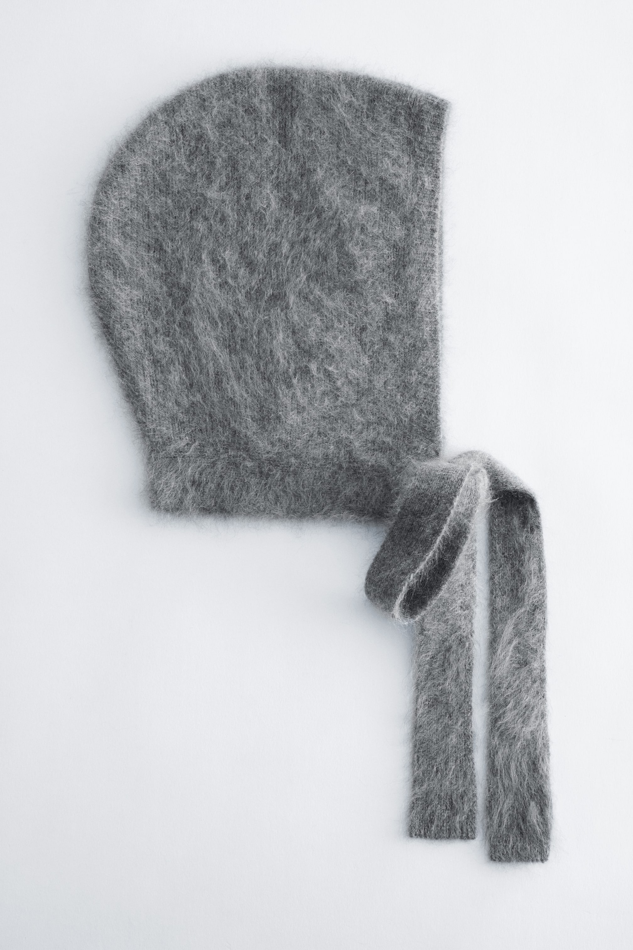 View larger image: A grey brushed cashmere bonnet is shown in side profile, highlighting its fuzzy, textured knit and rounded hood structure. Two long, flat-knit ties extend downwards from the front corner.