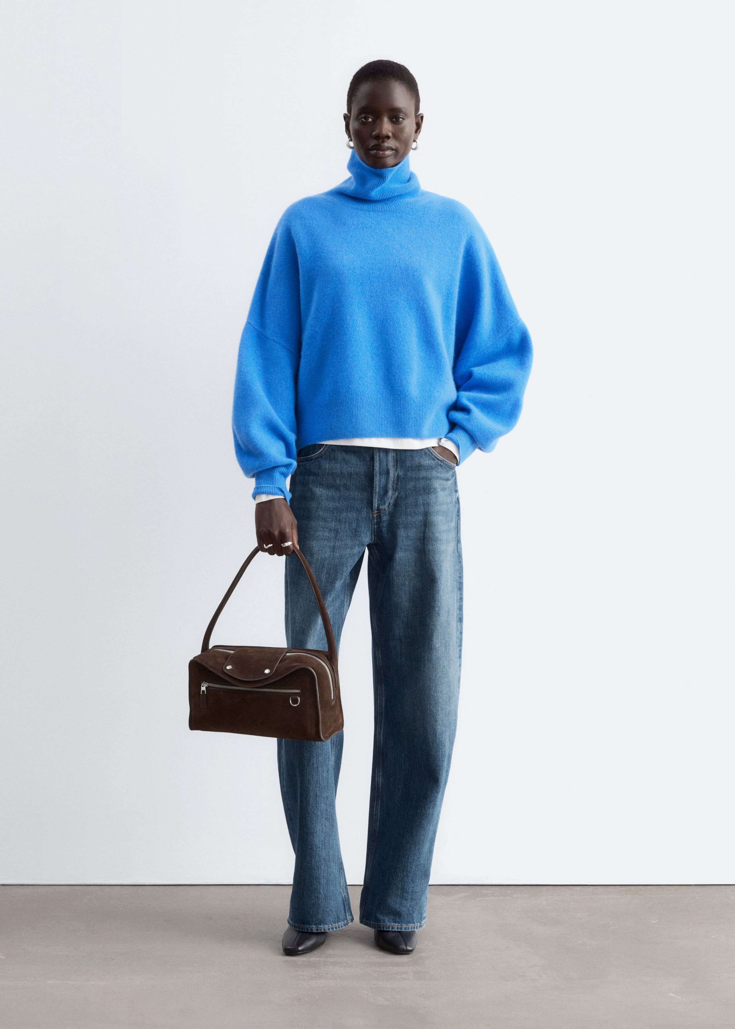An oversized, bright blue knitted jumper featuring a folded roll neck and dropped armholes is being worn by the model. The jumper is layered over a white top and styled with blue jeans and a brown suede handbag.
