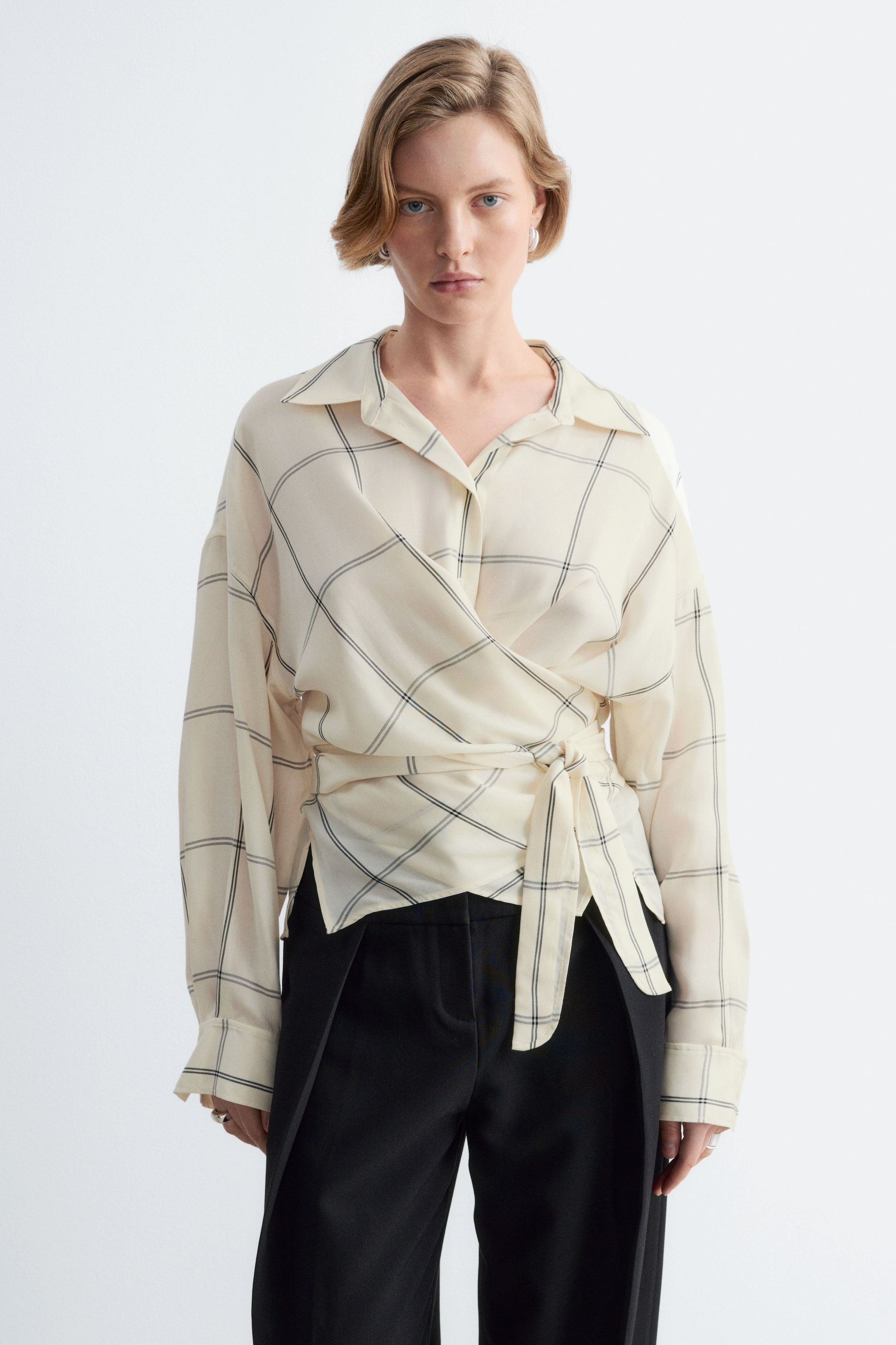 Agrandir l'image: Une chemise en coton couleur crème, à motif quadrillé, est portée par le mannequin. Elle présente une coupe cache-cœur nouée à la taille, un col classique et des manches longues. La chemise est associée à un pantalon noir taille haute.