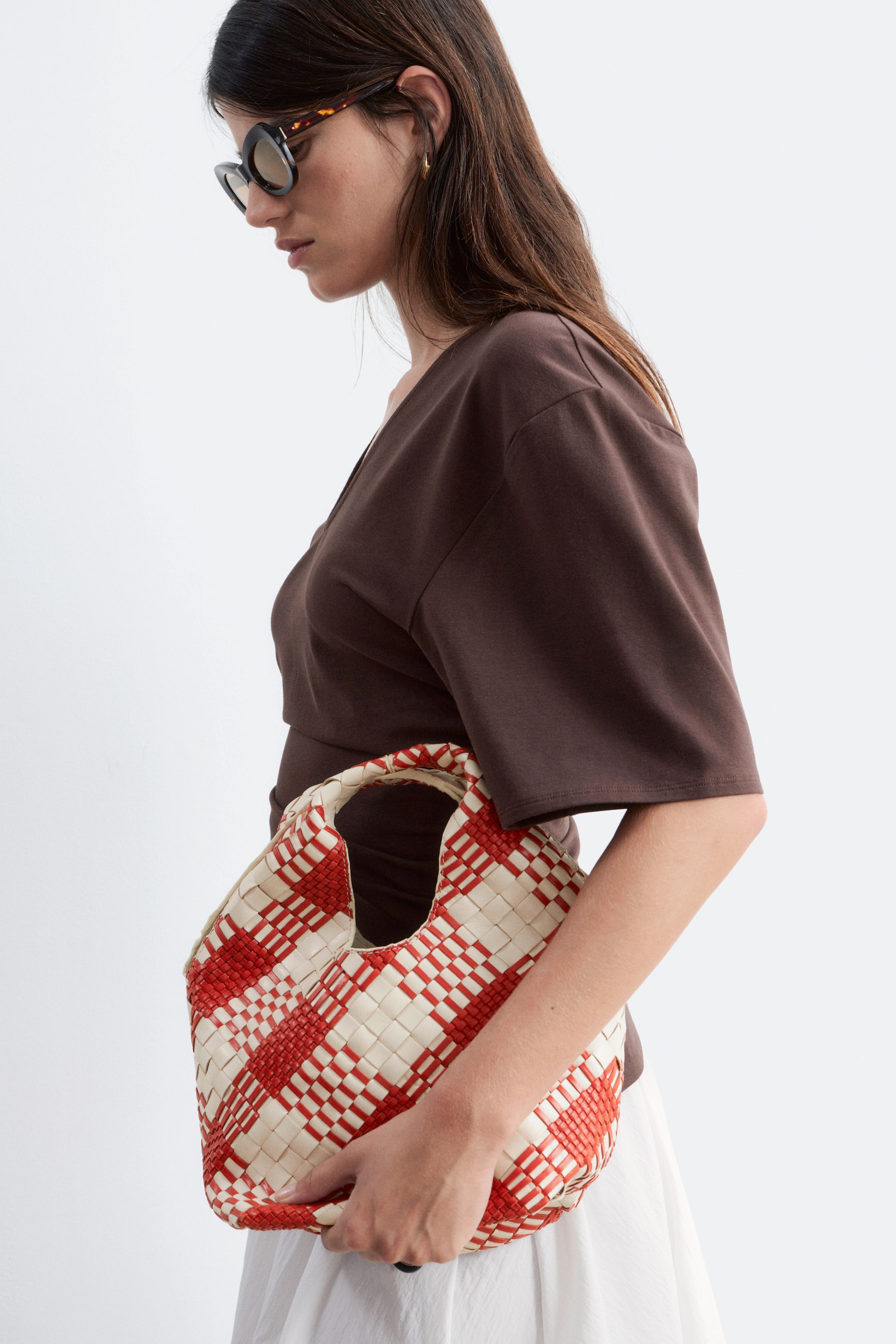 Visa större bild: Den vävda väskan visar ett rött och krämfärgat schackrutigt mönster, med ett integrerat topphandtag och en mjuk, rundad form. En hand håller väskan från en något vinklad sidovy, vilket framhäver dess struktur.