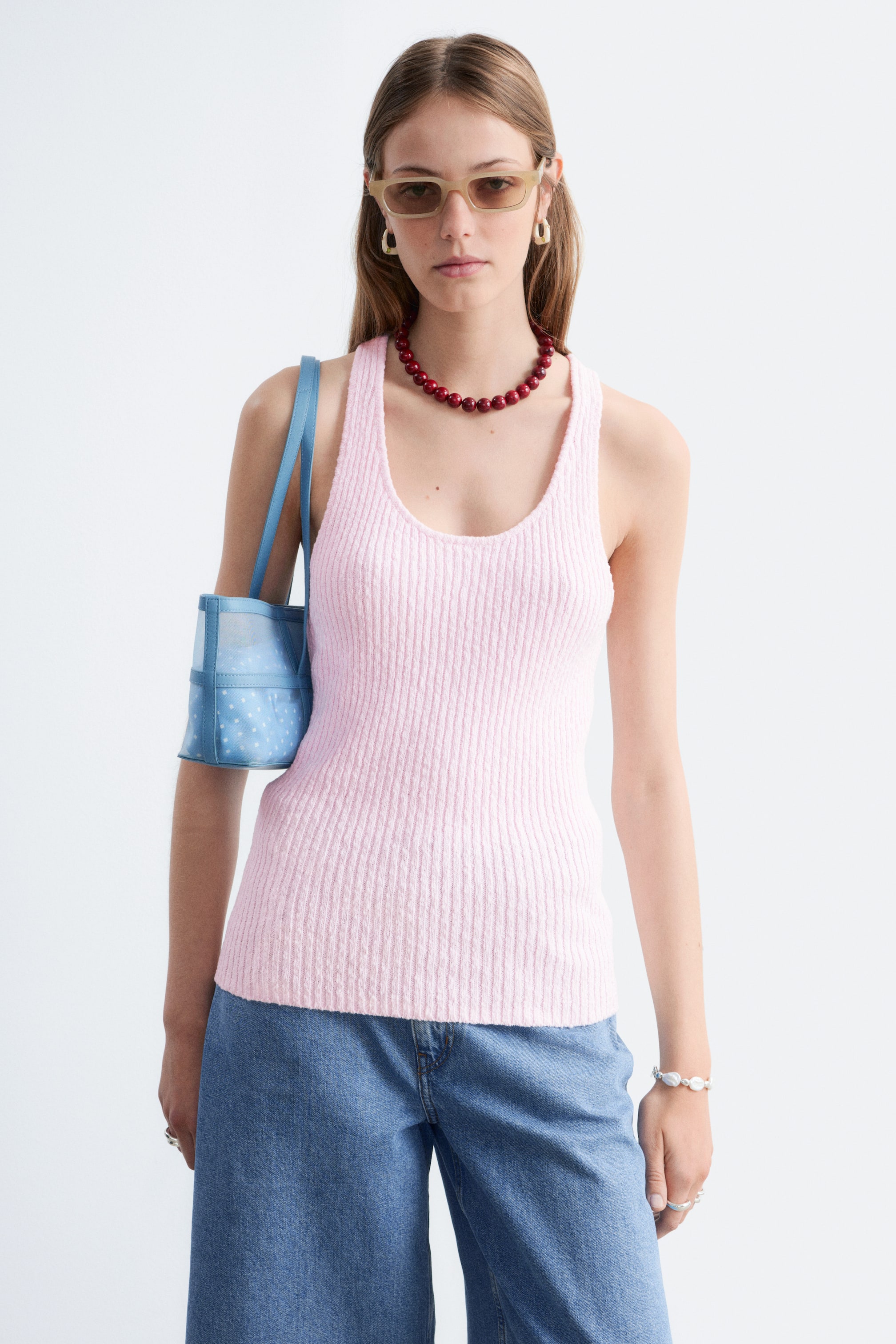 Grotere afbeelding bekijken: Roze geribbelde tanktop met een ronde hals en een aansluitende pasvorm, die tot aan de taille reikt. Het wordt gedragen onder een rode kralenketting en over een blauwe jeans.