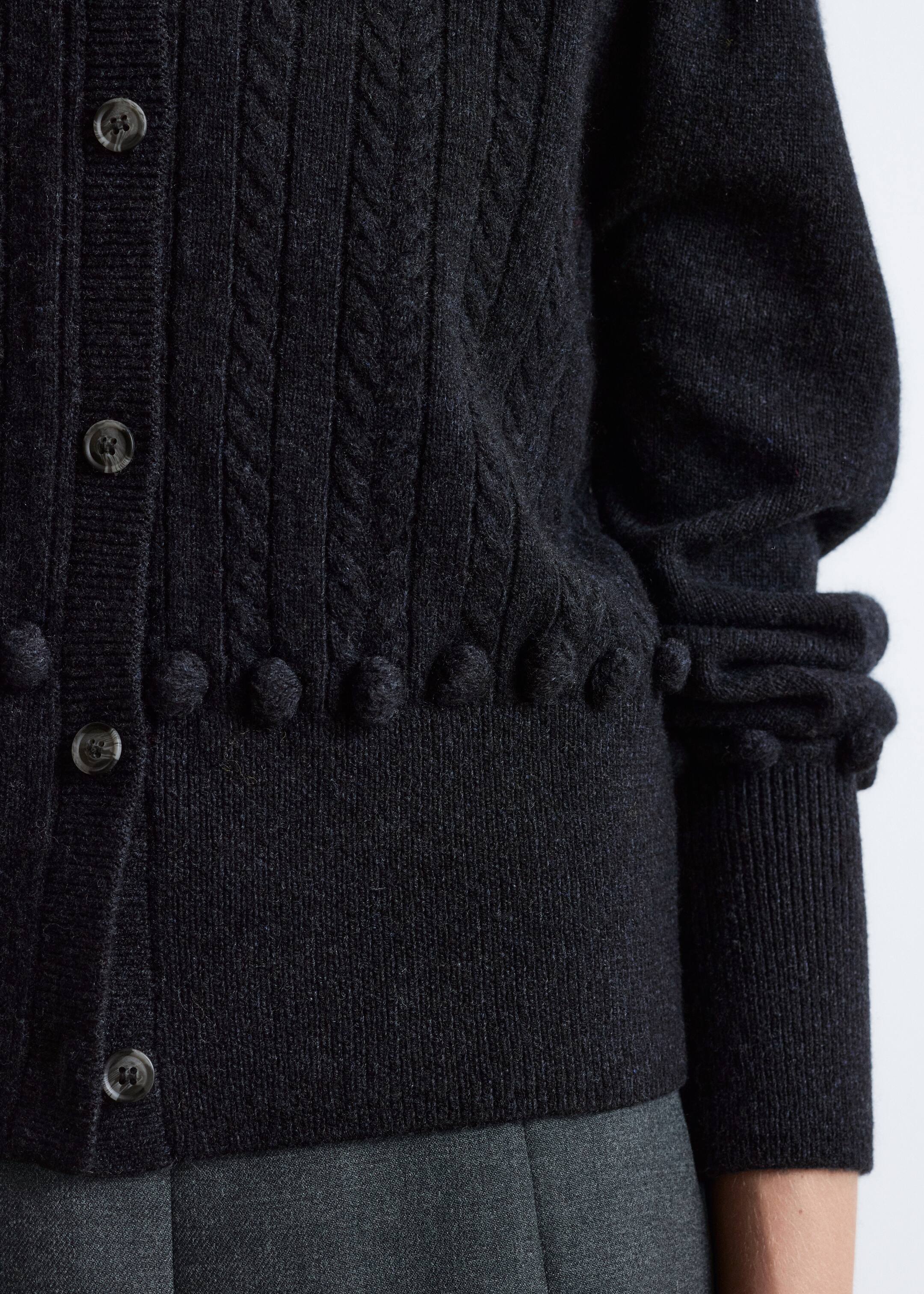 Ingrandisci l'immagine: Un cardigan grigio scuro viene mostrato da una prospettiva angolata, caratterizzato da un motivo a trecce sul corpo, una fila di dettagli a palline sopra l'orlo a coste, bottoni scuri lungo l'abbottonatura e una raccolta della manica prima di un polsino a coste.