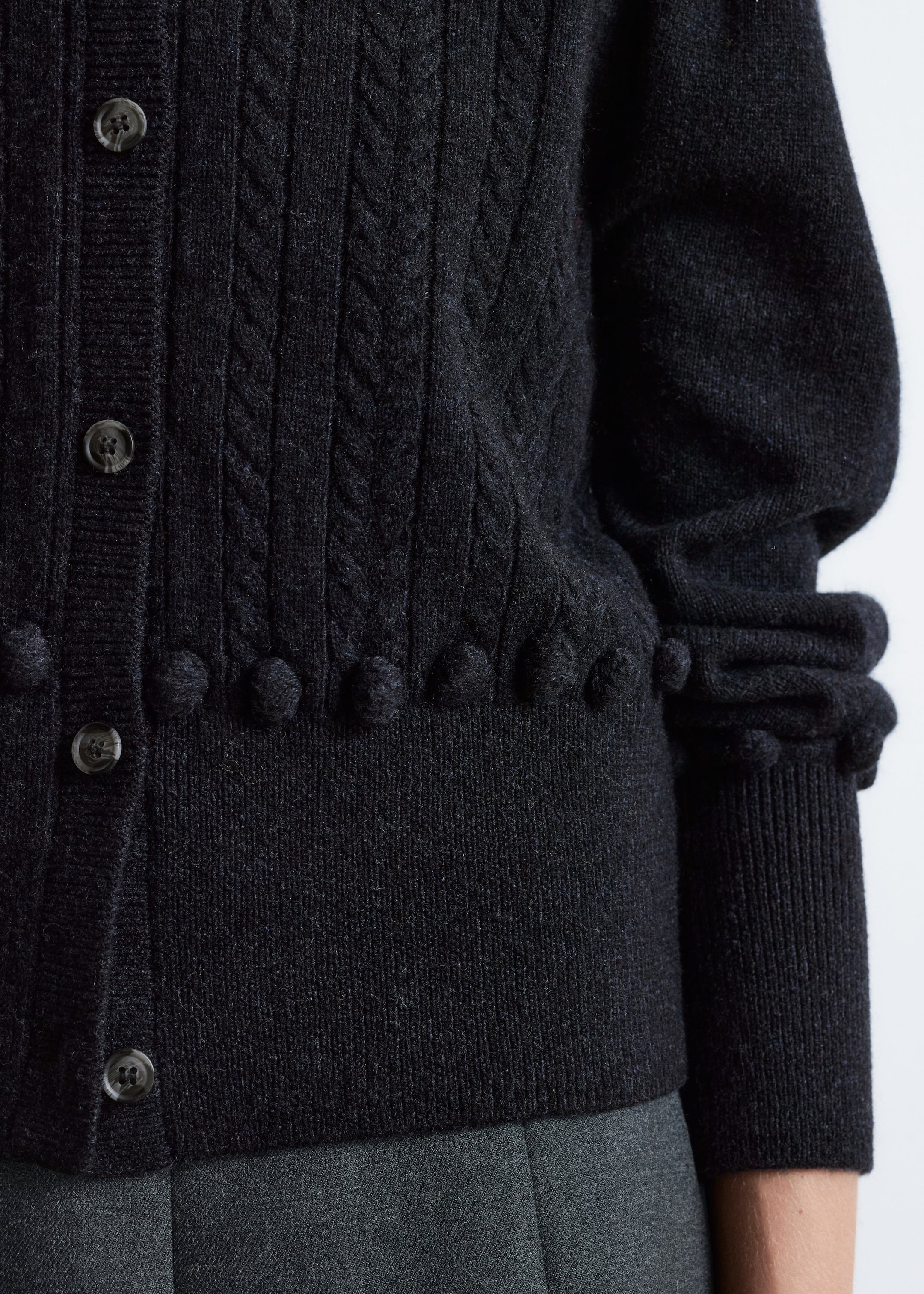 View larger image: A dark gray cardigan is being shown from an angled perspective, featuring a cable knit pattern on the body, a row of bobble detailing above the ribbed hem, dark buttons along the placket, and a sleeve gathering before a ribbed cuff.