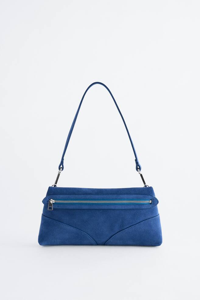 Un sac bandouliÃ¨re en daim bleu est prÃ©sentÃ© de face, affichant une silhouette rectangulaire avec des dÃ©tails de coutures incurvÃ©es en bas. Il est dotÃ© dâune poche zippÃ©e horizontale sur le devant avec des finitions de couleur argentÃ©e et est suspendu Ã  une fine bandouliÃ¨re assortie. - 1