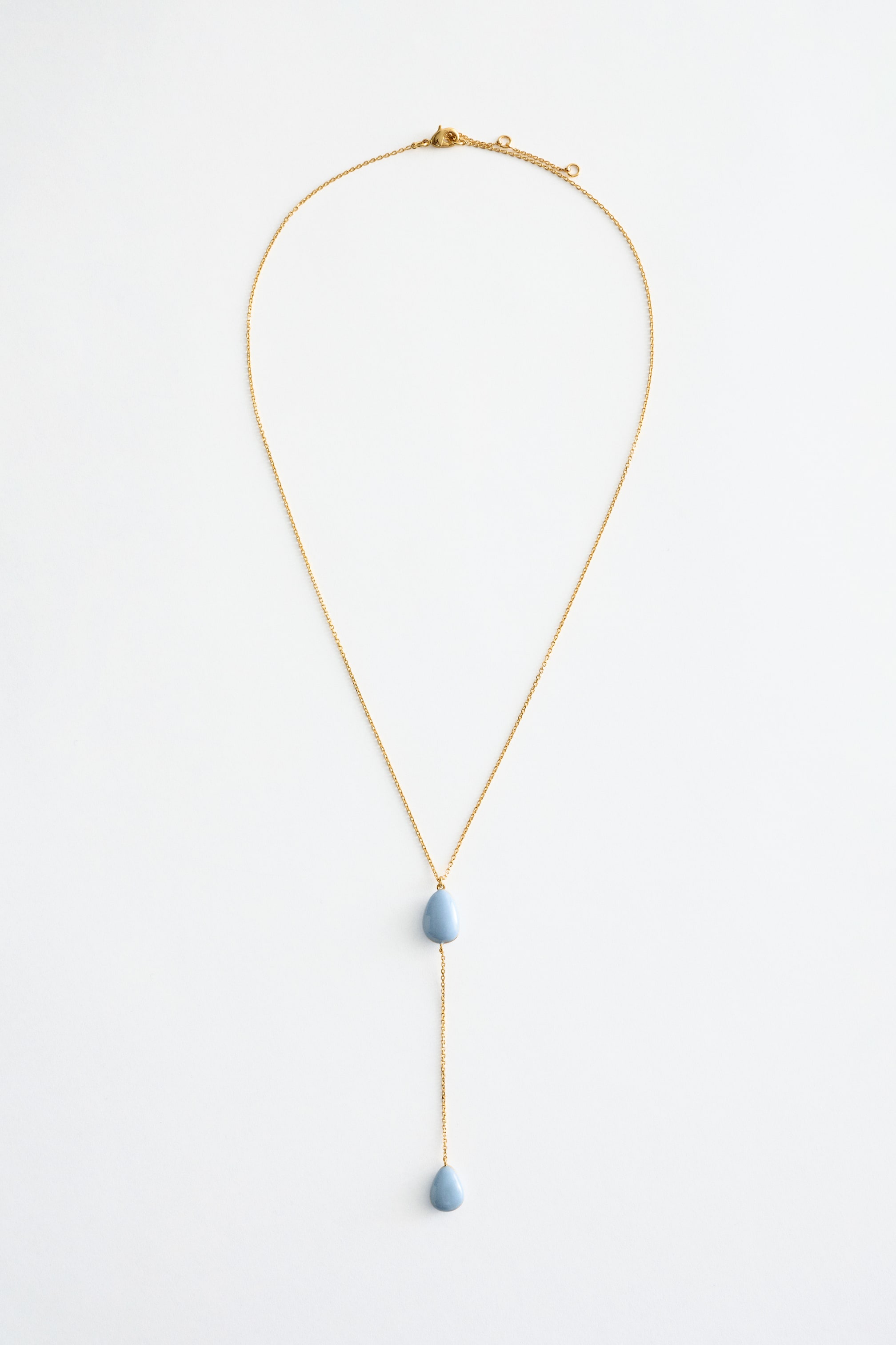 Grotere afbeelding bekijken: Een gepolijste messing ketting toont een lang, lariat-stijl silhouet, met twee lichtblauwe, druppelvormige hangers; één is gepositioneerd in het midden van de Y-splitsing, en de andere hangt aan het uiteinde van de ketting. Een verstelbare trigger sluiting is zichtbaar aan de bovenkant.