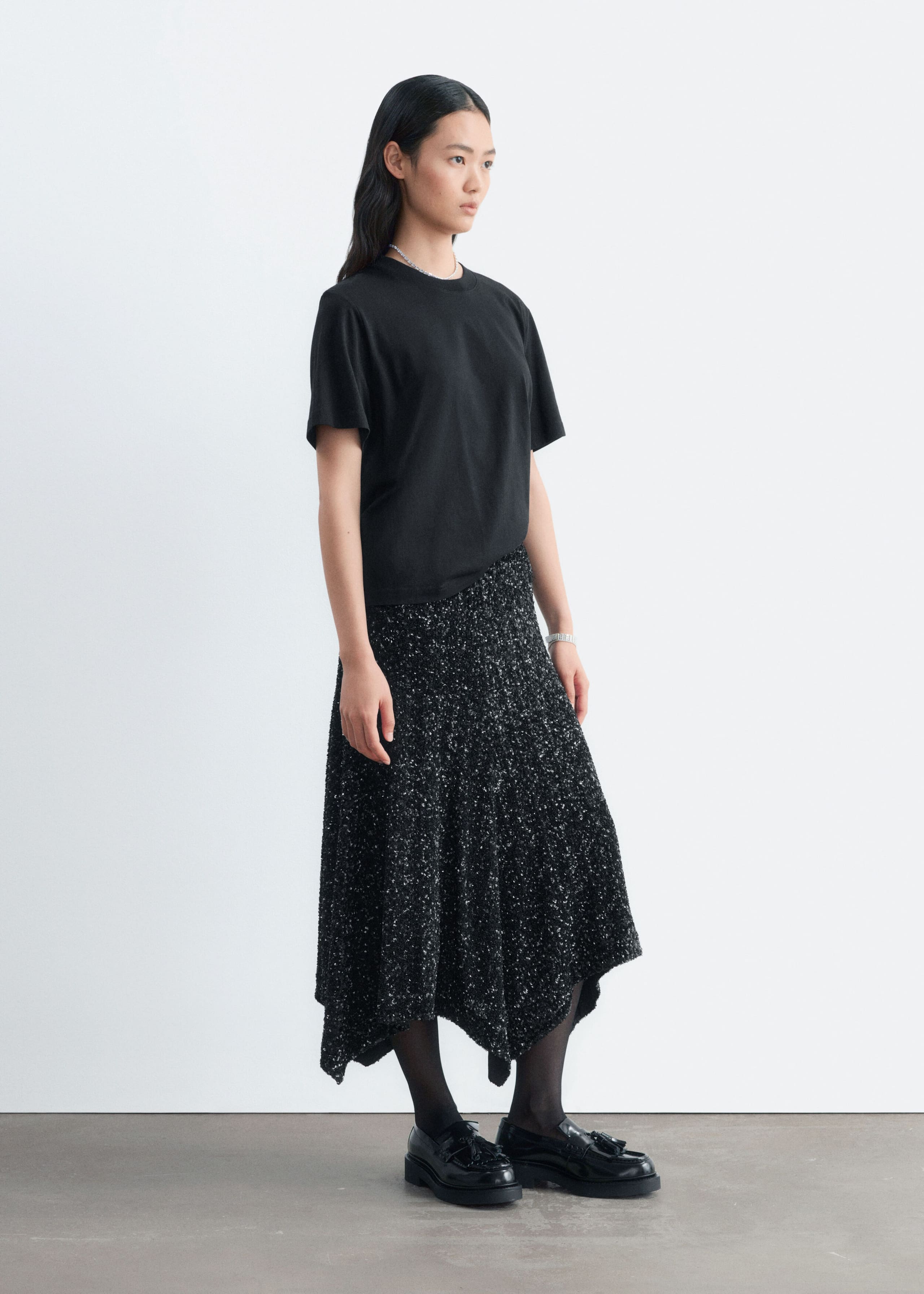 A black sequined mesh skirt with a textured appearance and an asymmetrical hemline is being worn by the model. The knee-length skirt is styled with a short-sleeve black T-shirt, sheer black tights, and black loafers.