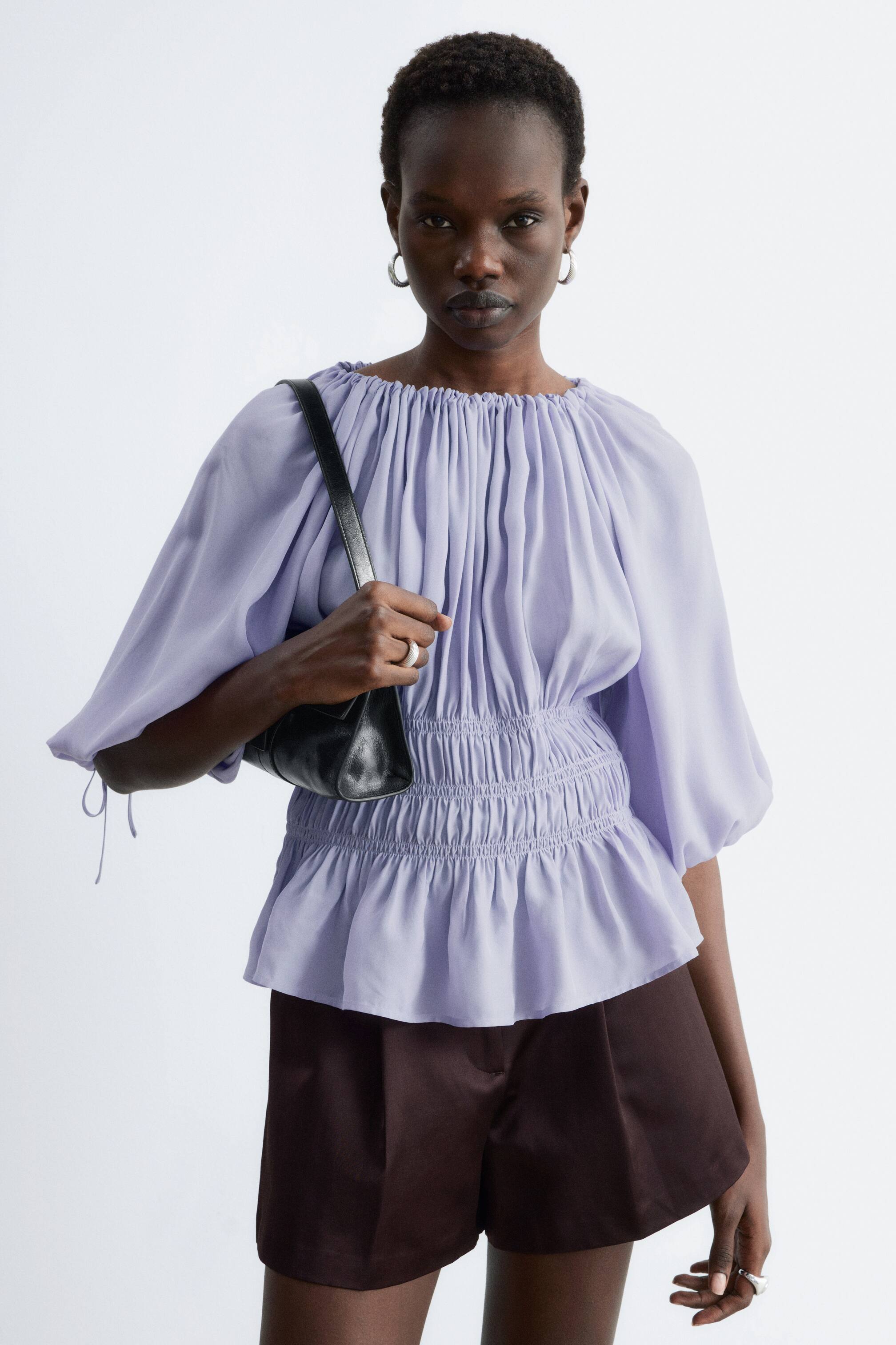 Größeres Bild ansehen: Die Model trägt eine lavendelfarbene Rüschenbluse mit einem gerafften Rundhalsausschnitt, voluminösen Ärmeln mit Bindedetails und einem gesmokten, elastischen Bund, der einen Schößchensaum bildet. Die Bluse wird mit braunen Shorts gestylt, und eine schwarze Umhängetasche wird getragen.