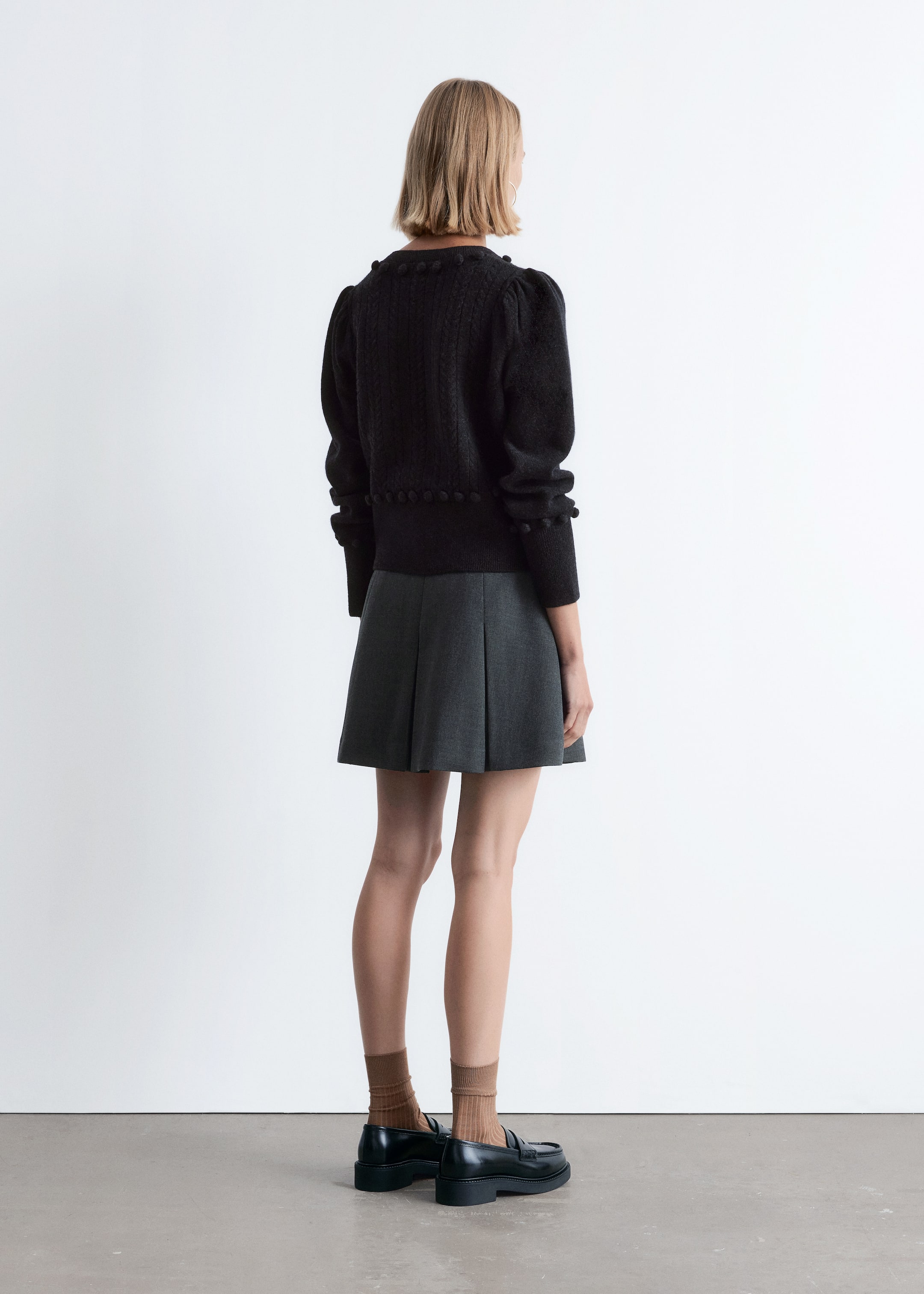 View larger image: A black cropped sweater is being shown from the back, featuring a textured knit pattern, puffed sleeves, and spherical embellishments along the hem, cuffs, and neckline. It is styled with a dark gray pleated mini skirt, light brown ribbed socks, and black chunky loafers.