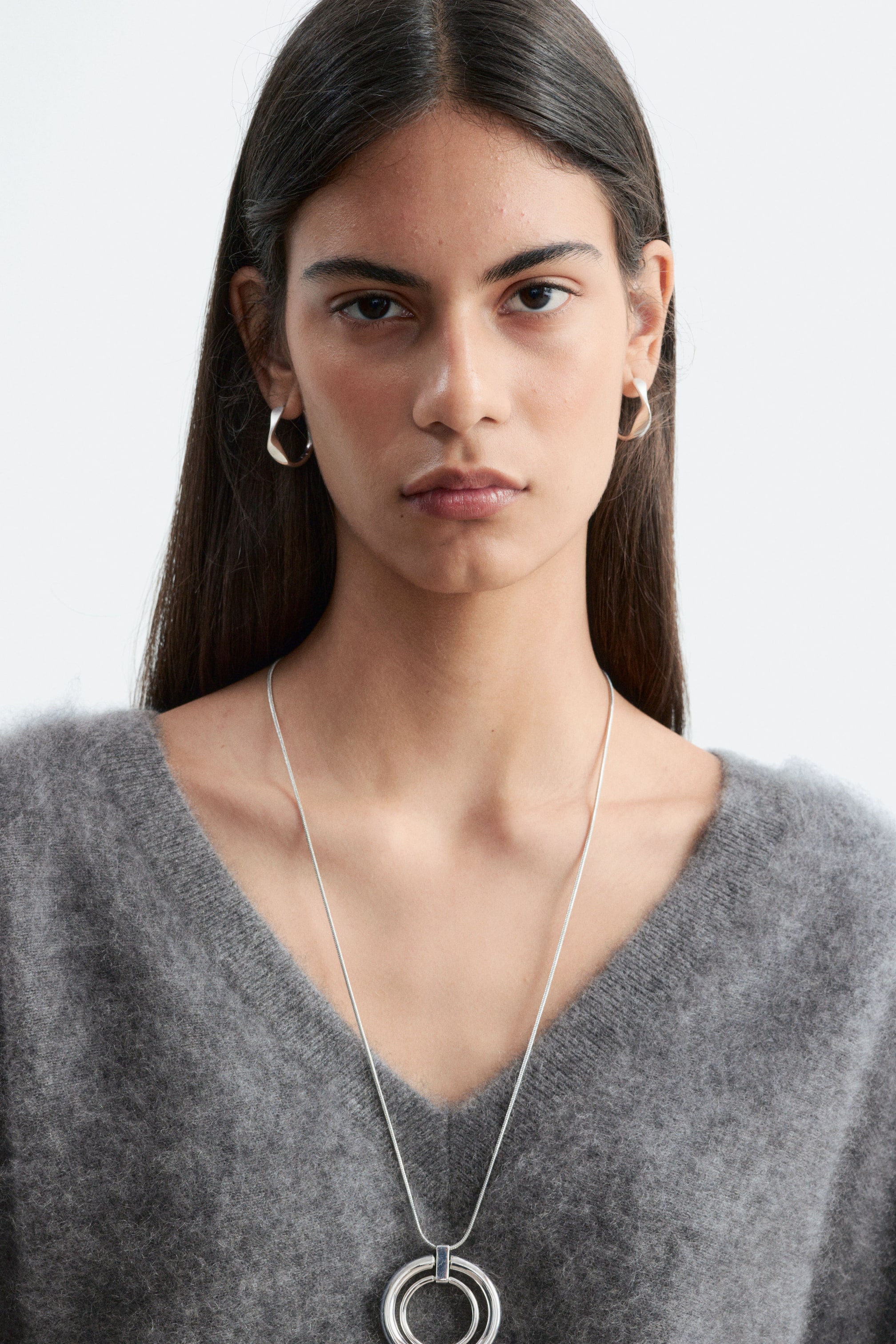 View larger image: A grey brushed cashmere V-neck jumper is being worn by the model, styled with a polished snake chain necklace featuring a double-circle pendant joined by a rectangular accent. The necklace rests against the model’s chest, showcasing its metallic finish against the soft texture of the jumper.