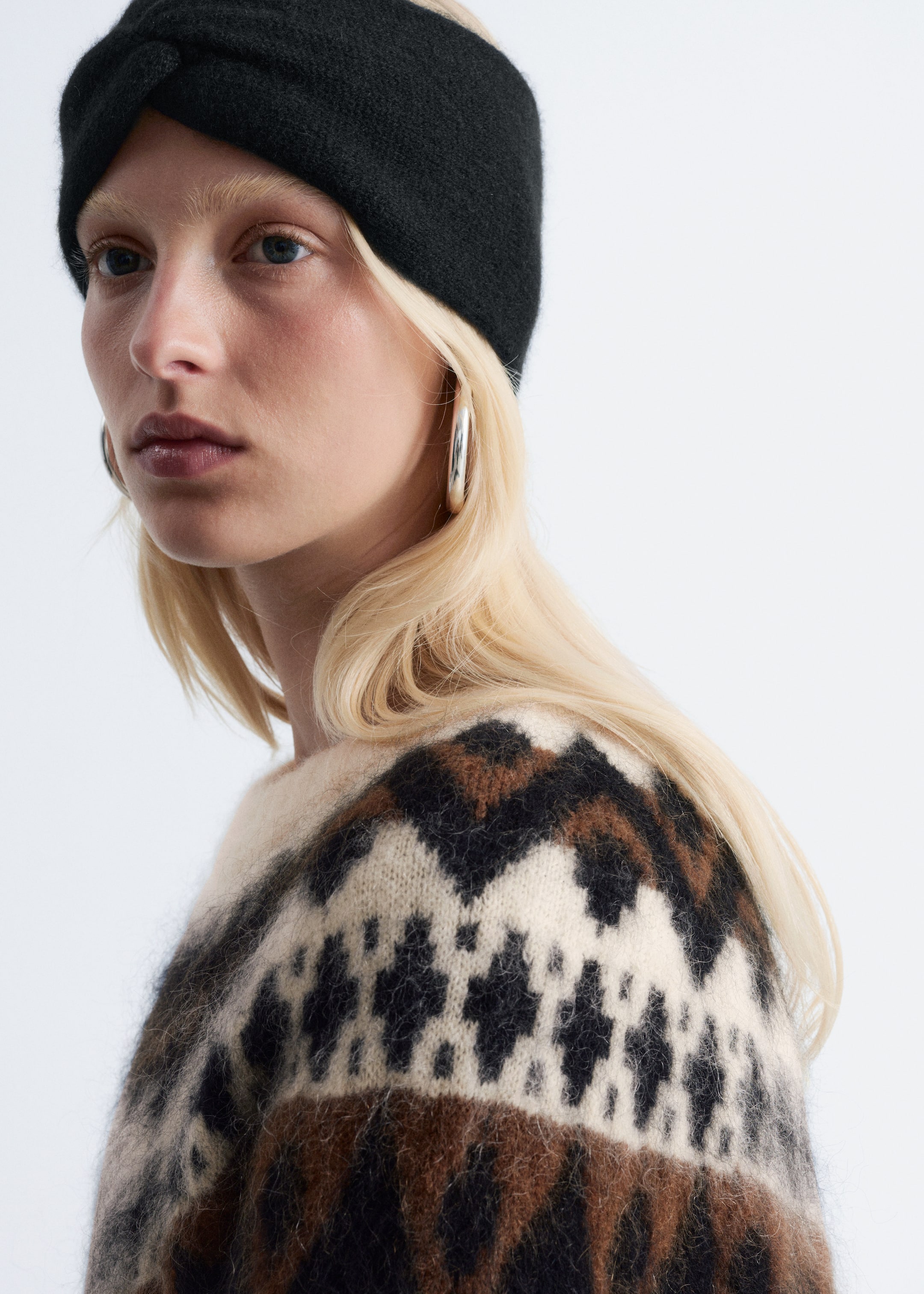 Agrandir l'image: Un bandeau en tricot noir présente un détail torsadé à l'avant, reposant sur le front. Un pull Fair Isle de couleur crème, marron et noir présente des motifs de losanges et de chevrons sur les épaules et le haut de la poitrine.