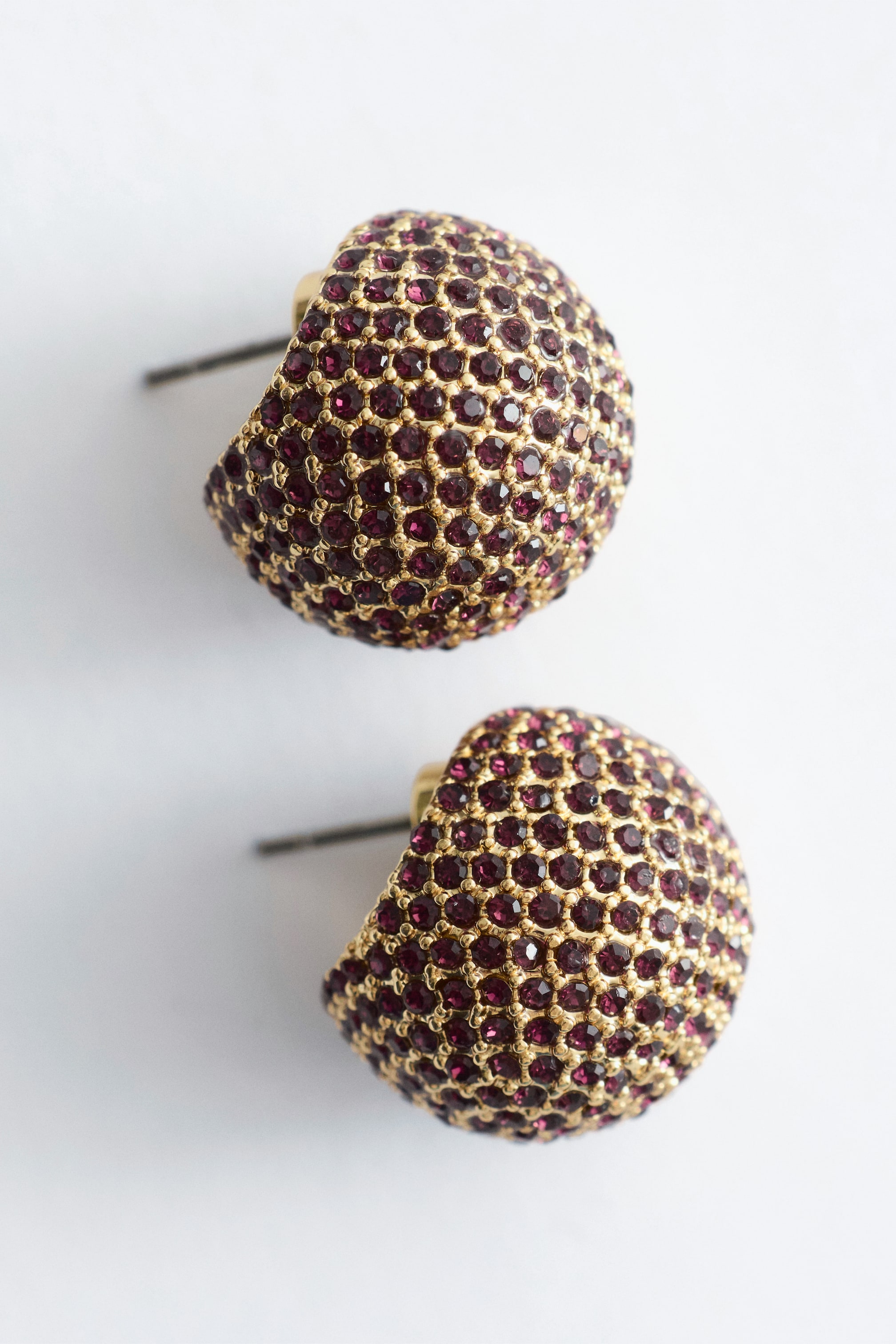 Visualizza immagine più grande: Un paio di orecchini a bottone a cupola è mostrato da un angolo leggermente inclinato, ciascun orecchino ricoperto da file di scintillanti strass color bordeaux incastonati su una superficie dorata. I perni metallici degli orecchini sono visibili.