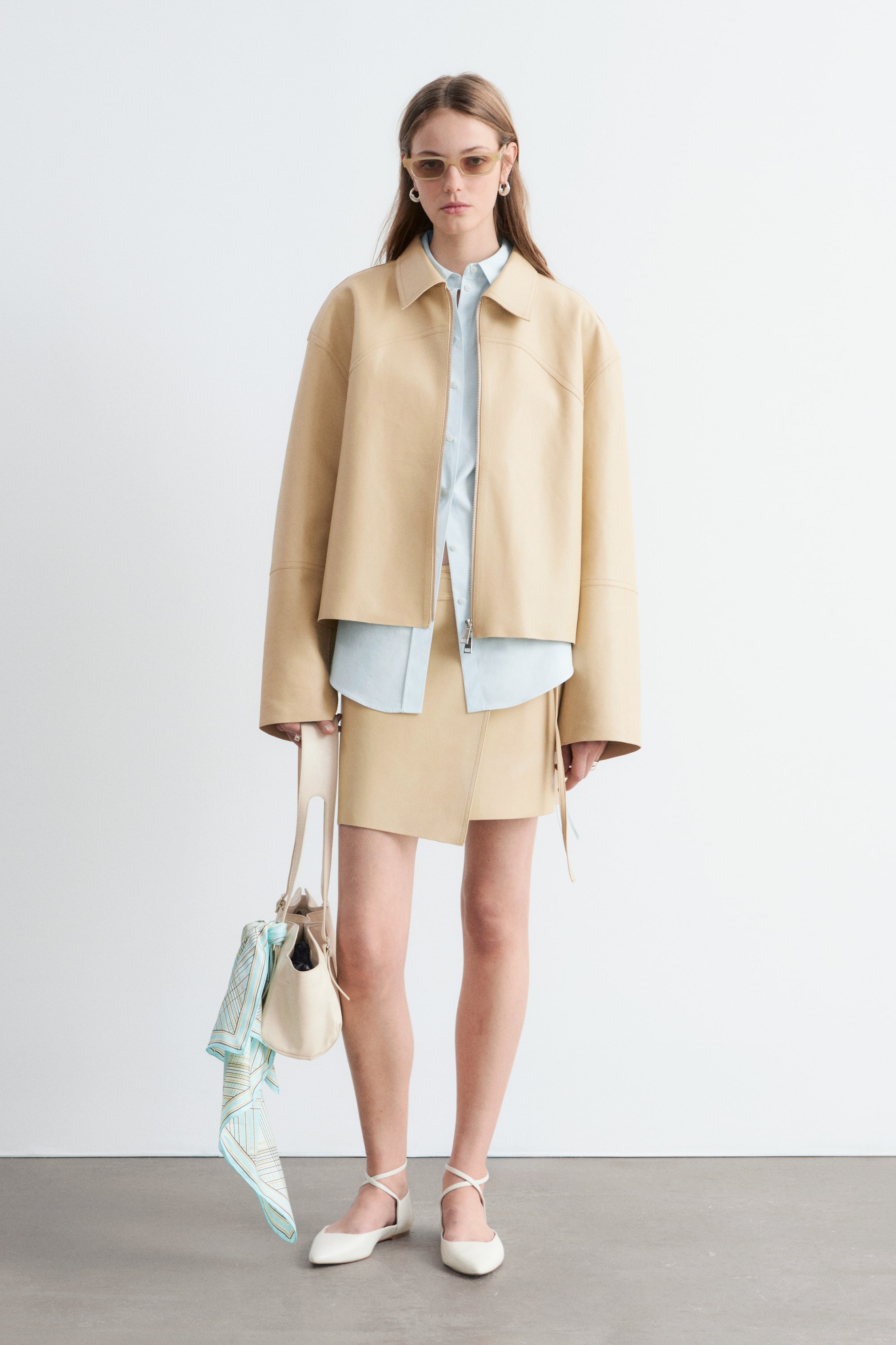 Nagyobb kép megtekintése: "Egy bézs színű, rövid bőrkabátot viselnek, amely hegyes gallérral és látható elülső cipzárral rendelkezik. Egy világoskék galléros ing fölé van rétegezve, amely laza szabást mutat, széles ujjakkal és varrási részletekkel a mellkasnál."