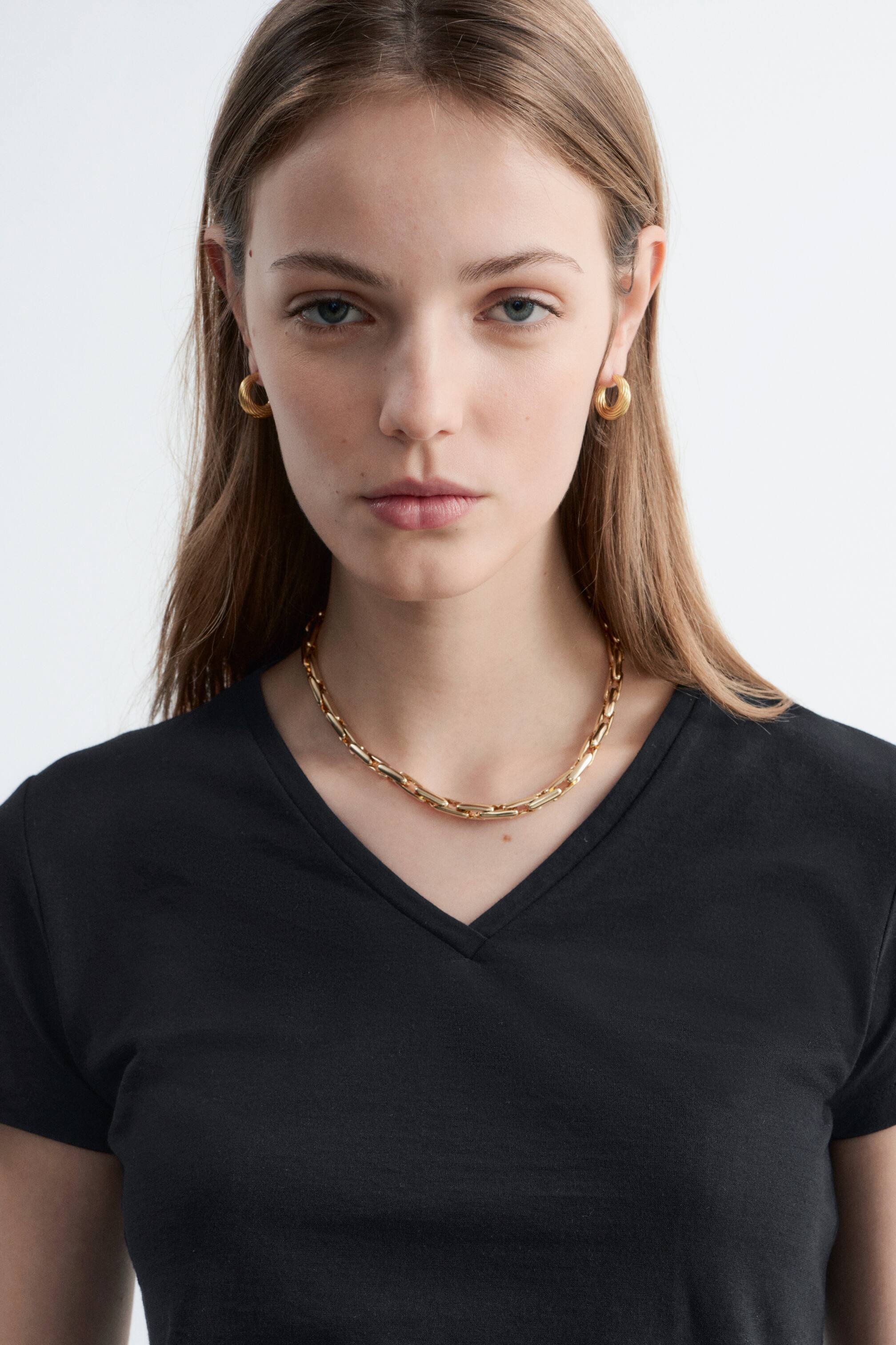 Se større billede: En sort V-hals t-shirt bæres af modellen. En poleret messing papirklipskæde bæres omkring modellens hals, hvilende over t-shirtens halsudskæring, og der bæres også guld hoop øreringe.