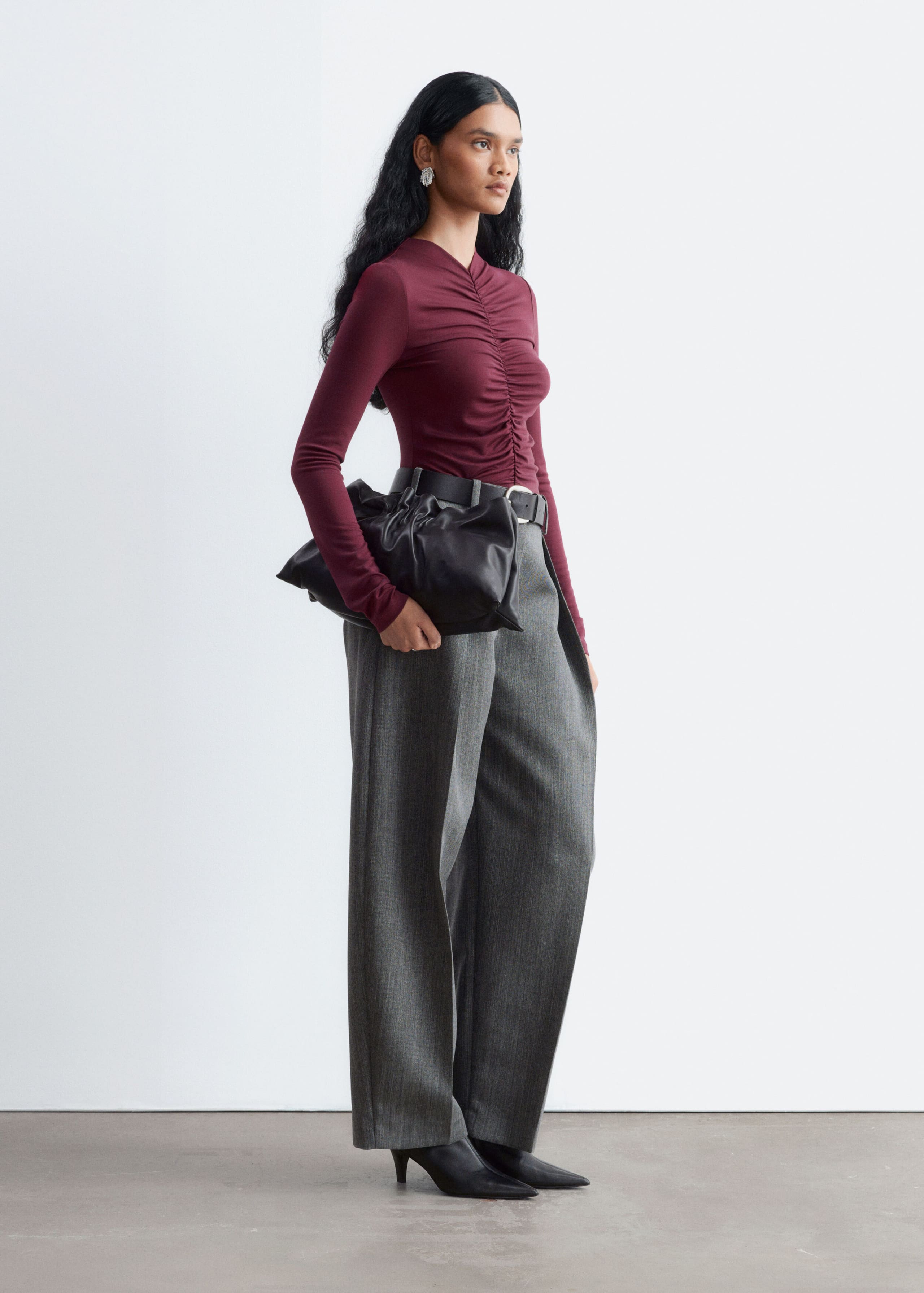 A burgundy long-sleeve top featuring gathered detailing along the center front seam is worn by the model, tucked into high-waisted gray wide-leg trousers with a black belt. The model is carrying a black leather clutch bag under one arm while standing in profile.