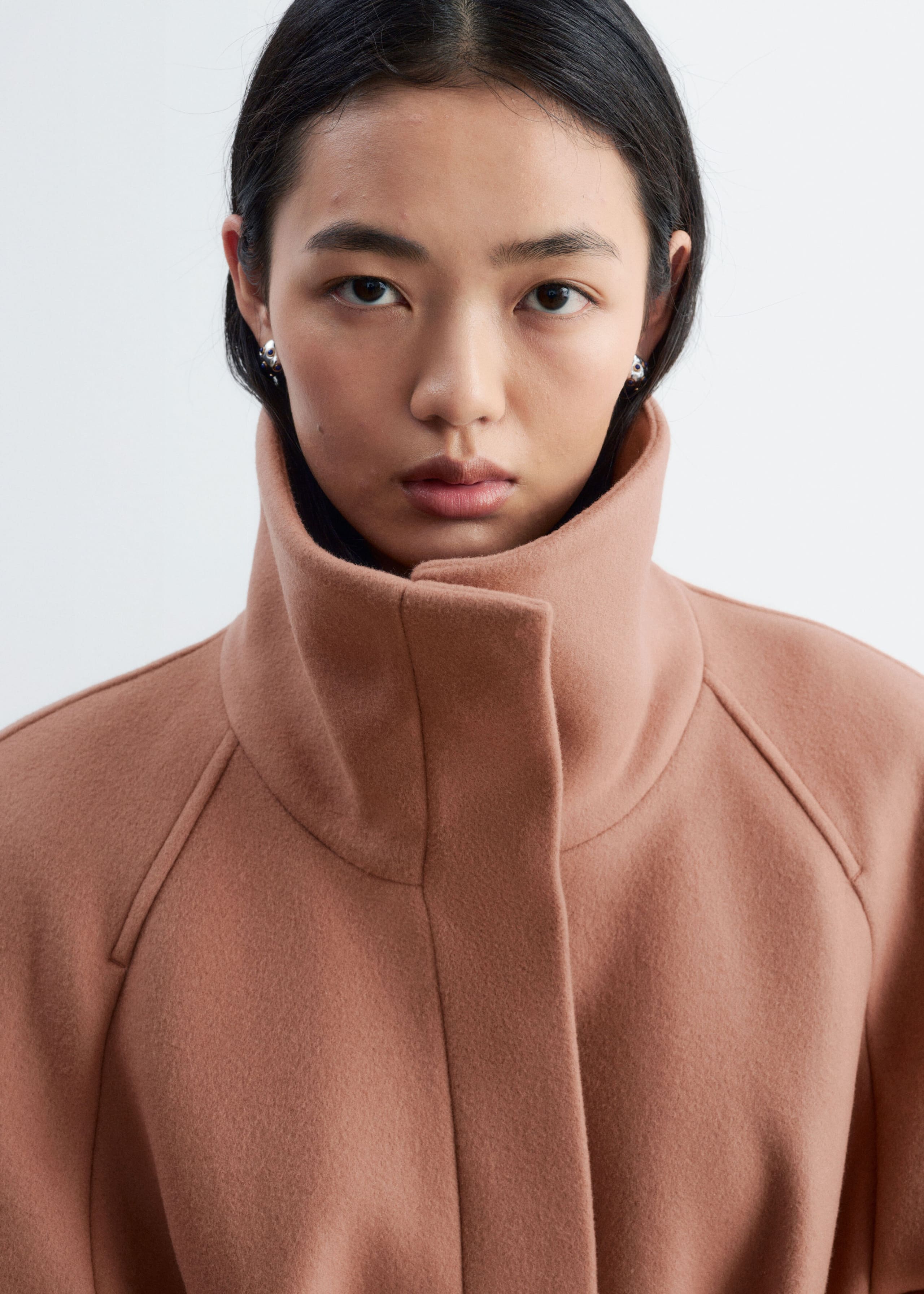 A dusty rose-coloured wool-blend coat is being worn by the model, showcasing a high, structured stand-up collar and a hidden front placket. The close-up view highlights the clean, minimalist design and raglan shoulder seams of the garment.