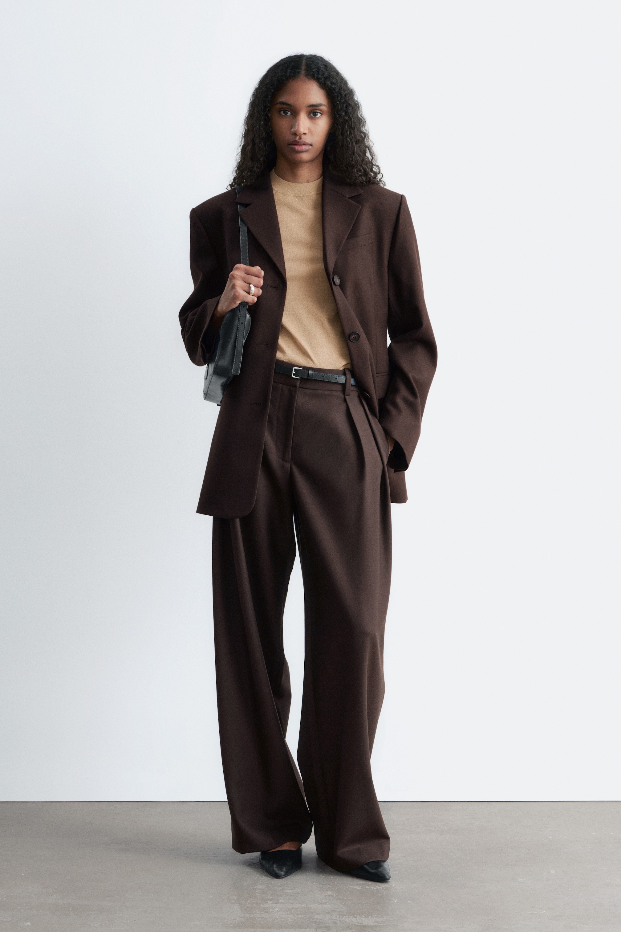 Näytä suurempi kuva: Mallin päällä on ruskea bleiseri, jossa on rakenteelliset olkapäät, kapeneva vyötärö, läppätaskut ja napitettu etuosa, kerrostettuna beigeen toppiin. Ruskeat leveälahkeiset housut, joissa on etulaskokset ja normaali vyötärö, on myös puettu, yhdistettynä mustaan vyöhön, samalla kun mallin käsi on asetettu viistoon sivutaskuun ja musta olkalaukku on mukana.