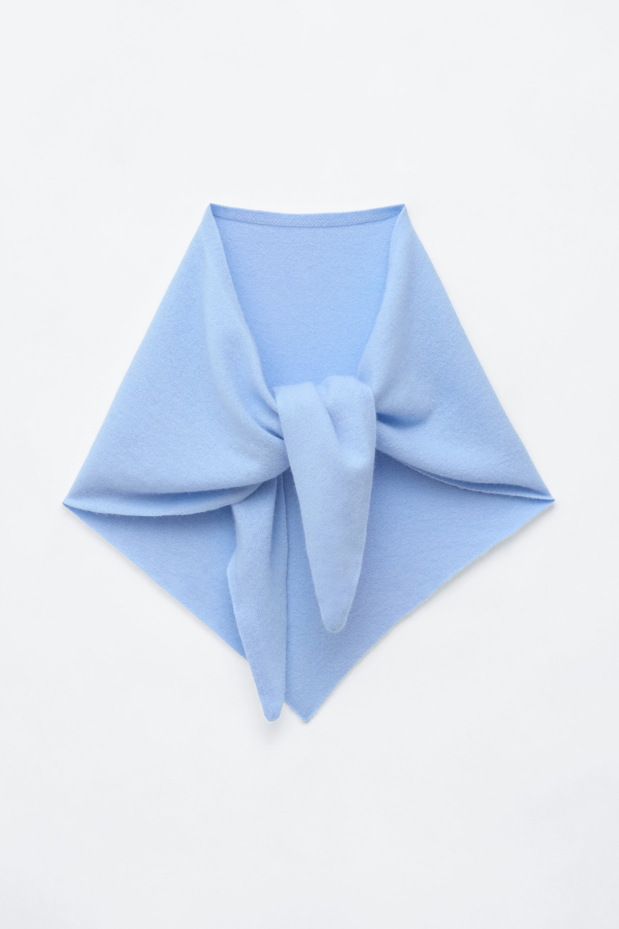 View larger image: A light blue knitted cashmere triangle scarf is draped in a front view, featuring a loose knot in the centre. The soft-textured fabric hangs in a symmetrical, diamond-like shape with the pointed corner pointing downwards.