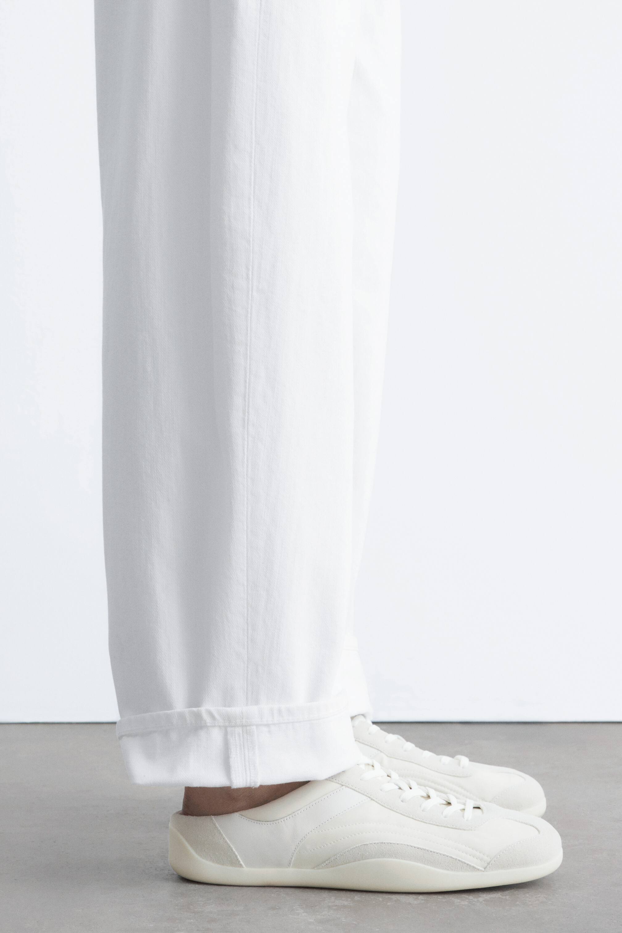 Se større billede: Off-white slip-on sneakers bæres af modellen, med en rund tå, en flad sål og et design, der kombinerer glatte og ruskindslignende paneler med synlige snørebånd. Hvide bukser med brede ben, opsmøget ved anklen, styler sneakersene.