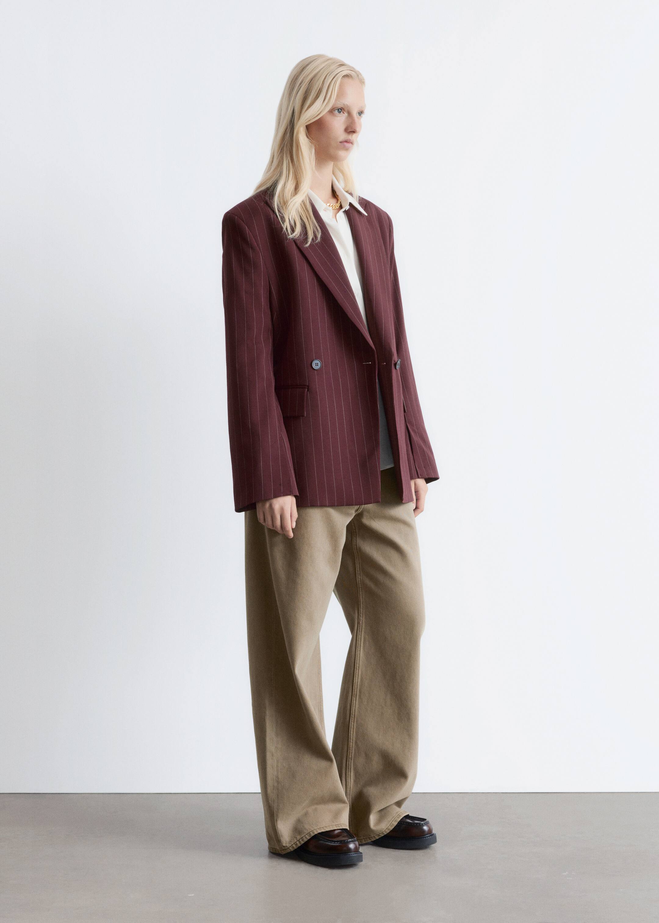 Agrandir l'image: Un blazer bordeaux à rayures fines et à double boutonnage, avec une double rangée de boutons sombres, des revers en pointe, des épaules structurées et des poches à rabat à l'avant, est porté ouvert par le mannequin. Il est superposé sur une chemise blanche et associé à un pantalon kaki à jambes larges.