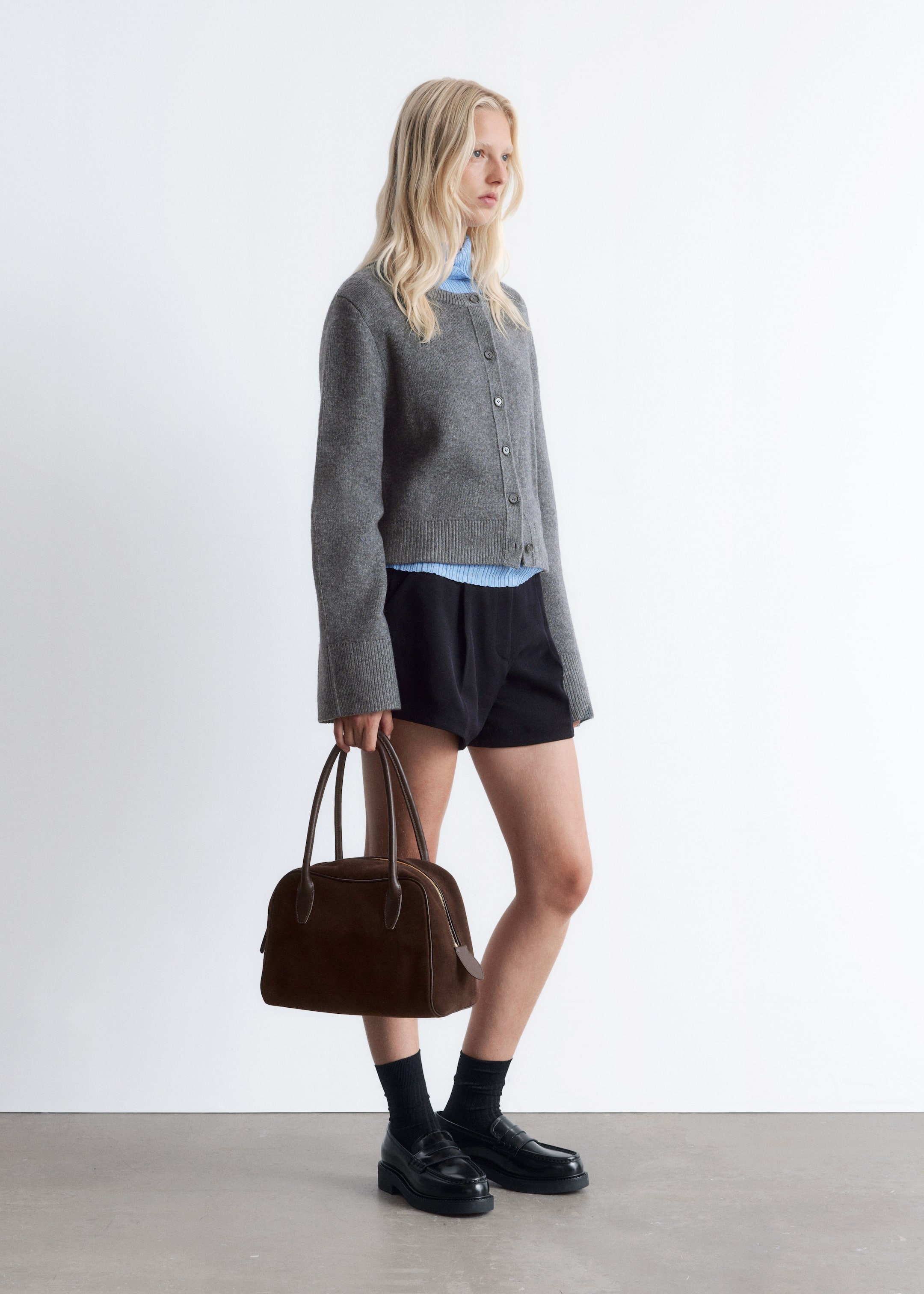 Vis større bilde: En grå cardigan med rund hals bæres, kneppet opp, lagd over en lyseblå ribbestrikket mock-neck skjorte og svarte løstsittende shorts, presentert fra en sidevinkel. Antrekket kompletteres med svarte chunky loafers, svarte ribbestrikkede sokker, og en brun strukturert veske med topphåndtak og synlig glidelåslukking, som holdes.