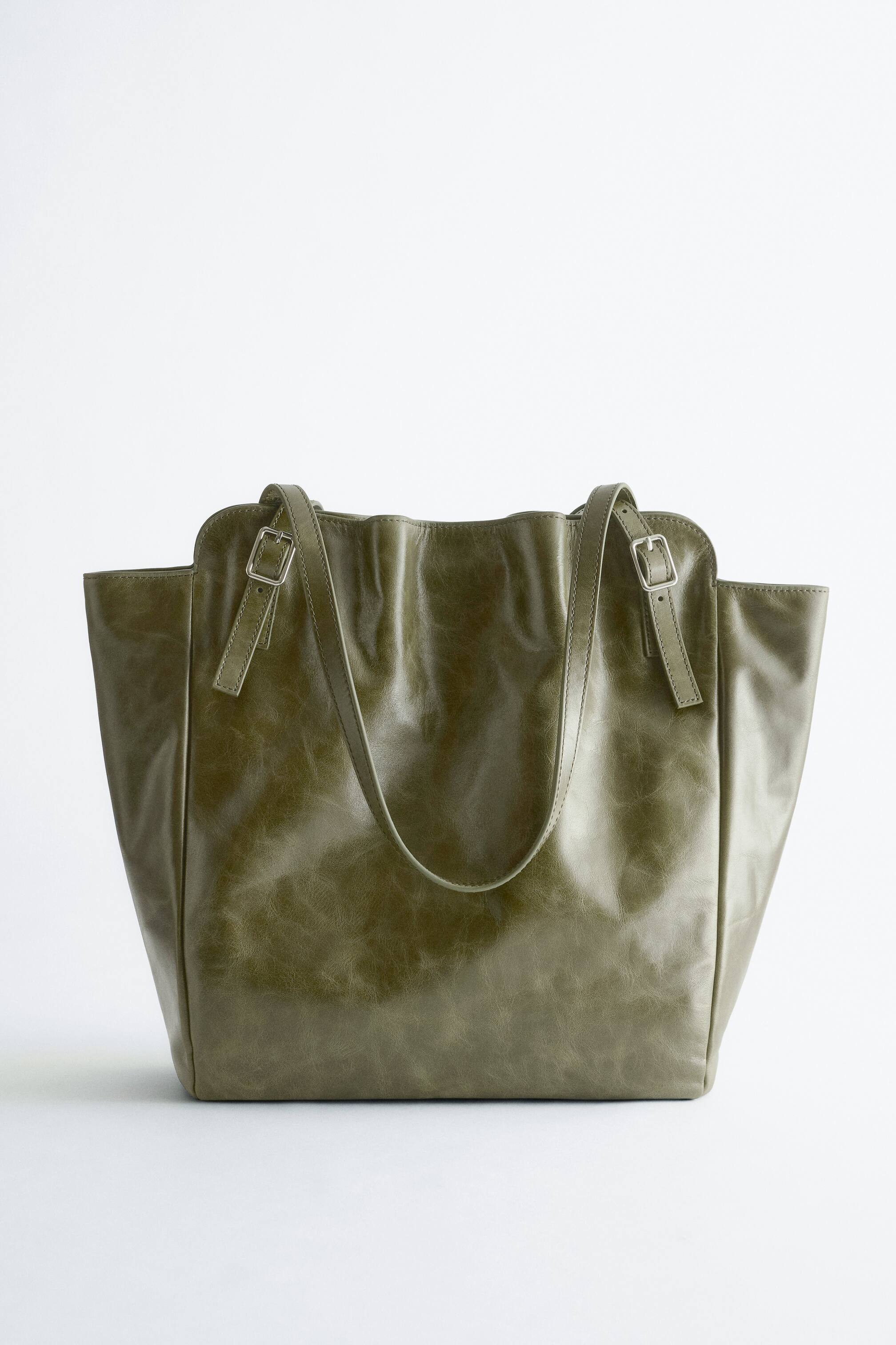 Agrandir l'image: Le sac cabas en cuir à boucle présente sa face avant, confectionné en cuir vert olive avec une finition légèrement vieillie. Deux anses d'épaule émergent de l'ouverture supérieure, où deux petites boucles argentées resserrent les côtés supérieurs du sac.
