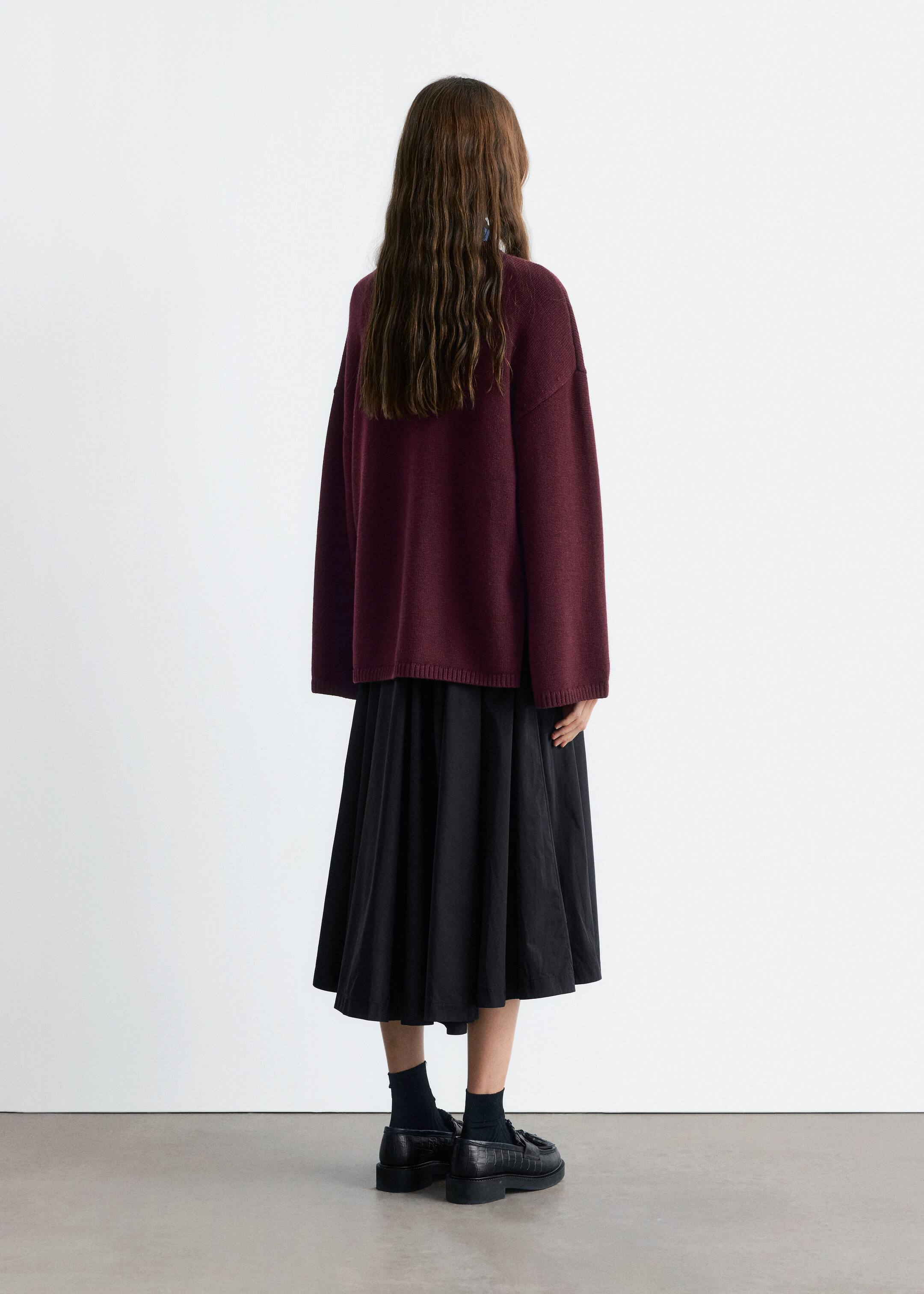 Agrandir l'image: Un pull oversize bordeaux, avec de larges manches longues dotées de poignets côtelés et un ourlet côtelé, est présenté de dos. Il est superposé à une jupe midi plissée noire, le look complet étant complété par des chaussettes montantes foncées et des mocassins noirs à embossage crocodile avec des semelles épaisses.