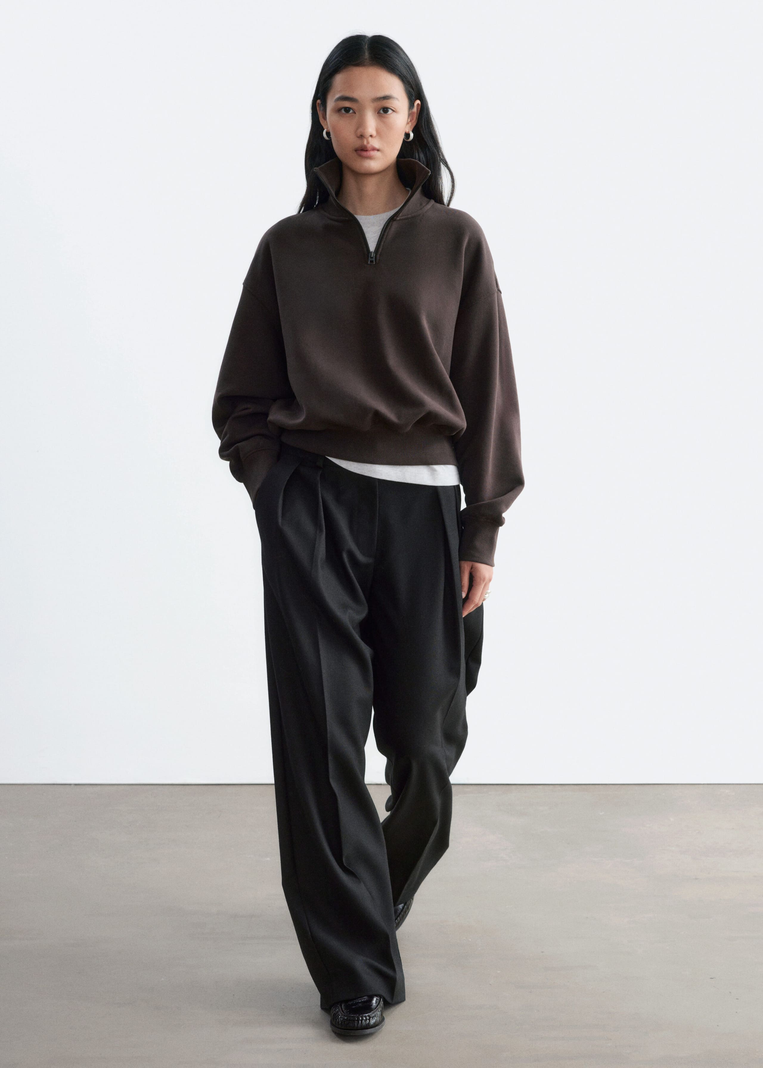 The model is wearing a dark brown cotton jersey sweatshirt with a half-zip collar and ribbed trims. The relaxed-fit sweatshirt is layered over a light grey T-shirt and paired with wide-leg black trousers.